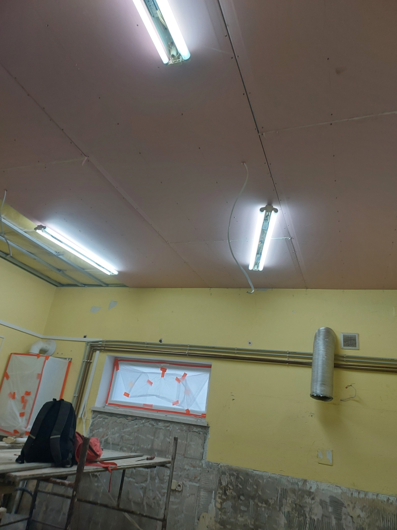 Płyty gipsowo-kartonowe mocowane do sufitu na metalowej konstrukcji; widoczne lampy fluorescencyjne, okno zabezpieczone folią malarską, fragment ściany z widocznymi śladami po pracach remontowych...