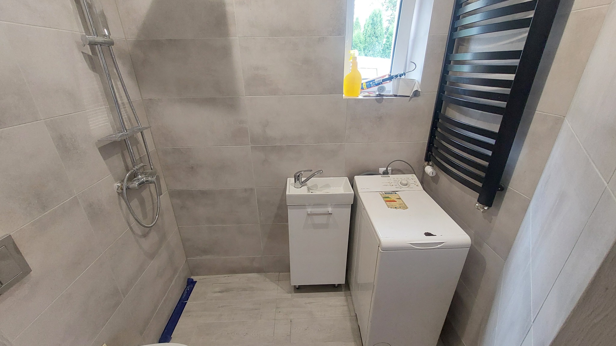 Jasna łazienka po remoncie: szare płytki, biała umywalka z szafką, pralka i czarny grzejnik drabinkowy. Nowoczesny design, schludne wykończenie.