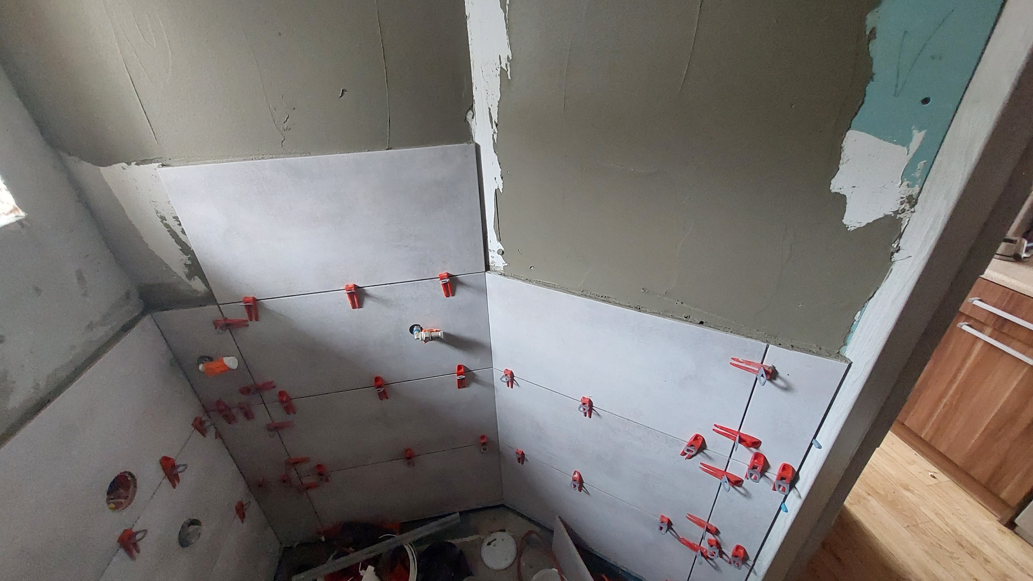 Ściany łazienki w trakcie układania jasnych płytek, widoczne krzyżyki dystansowe i fragmenty nieotynkowanych ścian. Ujęcie z dołu.