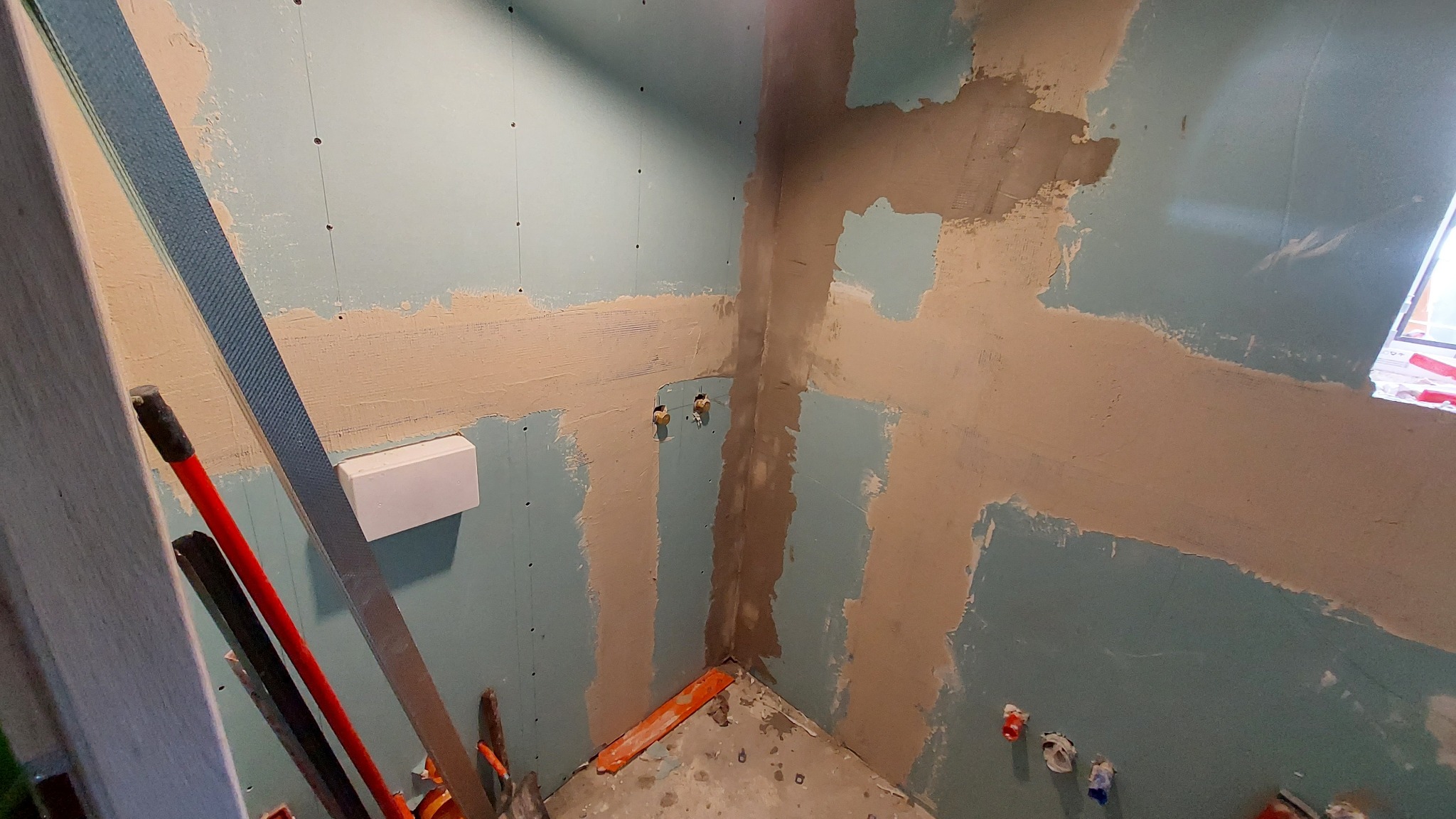 Narożnik łazienki w trakcie prac wykończeniowych: płyty gipsowe, częściowo położona gładź, widoczne instalacje wodne i narzędzia. Oświetlone światłem dziennym.