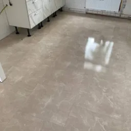 Beżowe płytki podłogowe z marmurkowym wzorem w kuchni, widoczne białe szafki kuchenne na czarnych nóżkach oraz grzejnik pod oknem.