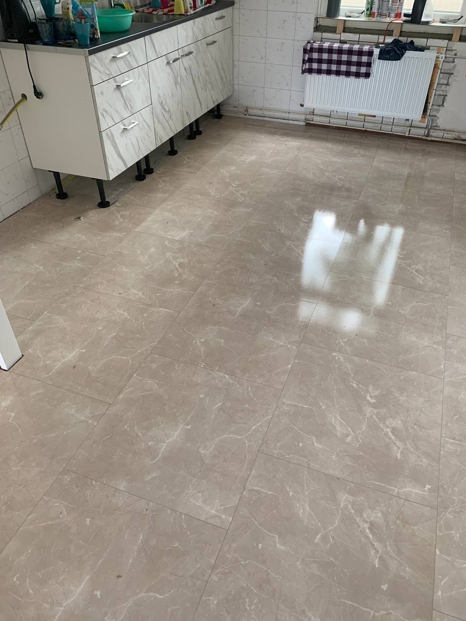 Beżowe płytki podłogowe z marmurkowym wzorem w kuchni, widoczne białe szafki kuchenne na czarnych nóżkach oraz grzejnik pod oknem.