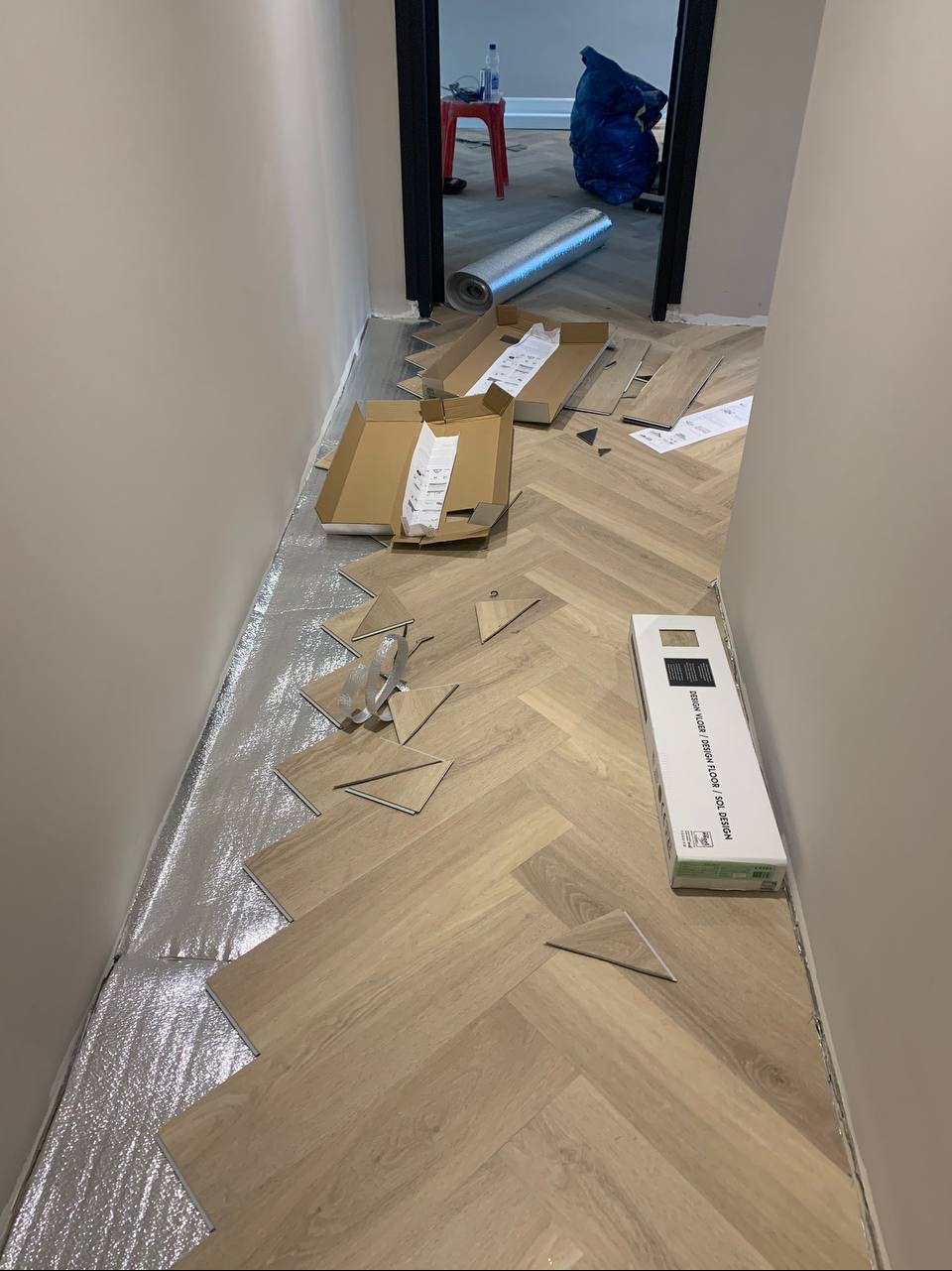 Układanie jasnych paneli podłogowych w jodełkę; widoczna folia izolacyjna, otwarte kartony z panelami i akcesoria montażowe w wąskim korytarzu.