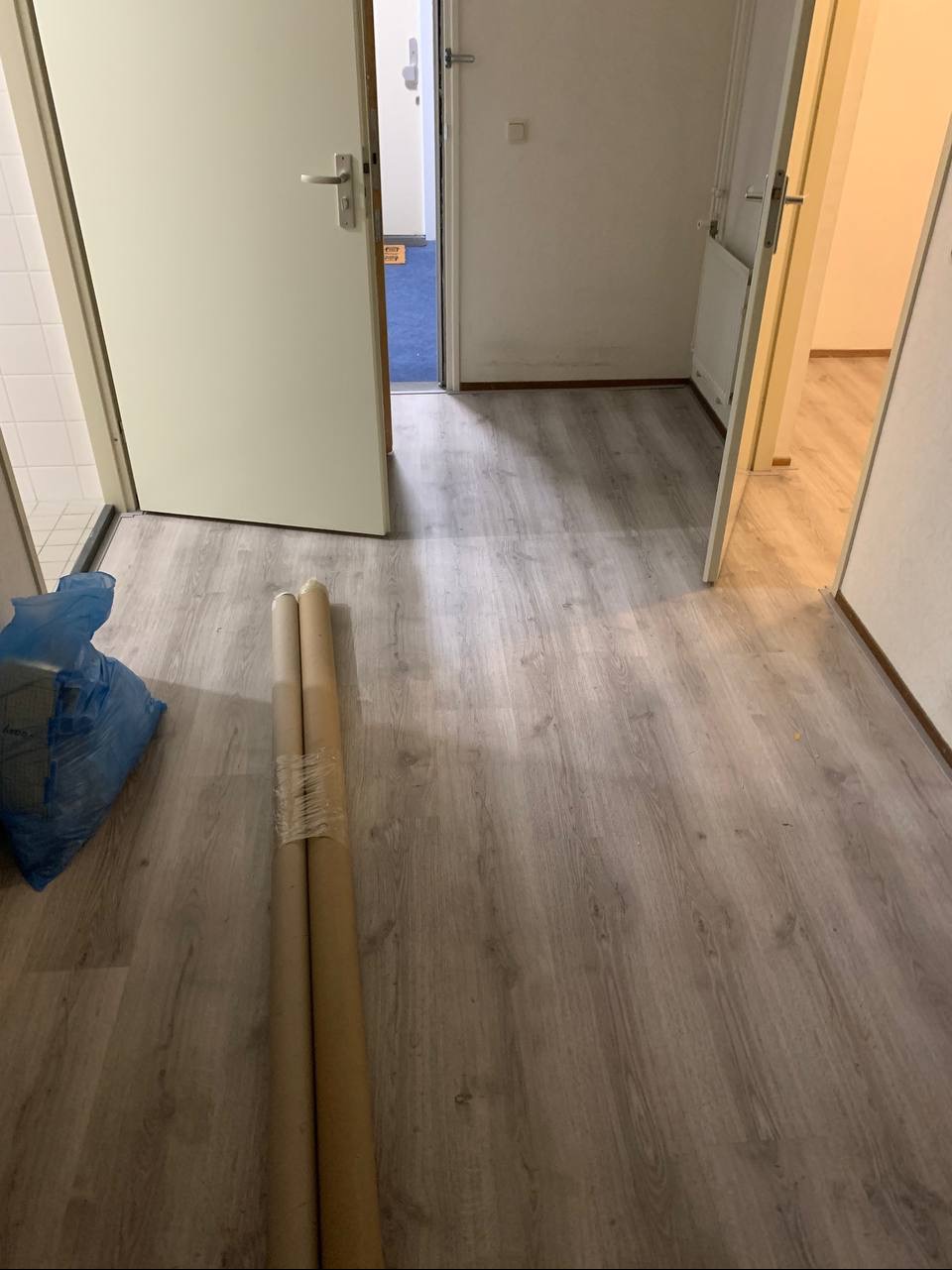 Nowo ułożone panele podłogowe w korytarzu, widoczne dwie zrolowane paczki paneli zabezpieczone folią i niebieski worek z odpadami.