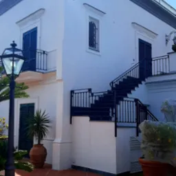 Dwupiętrowy budynek w stylu śródziemnomorskim z białymi ścianami, niebieskimi okiennicami i czarnymi balustradami, widok z tarasu z terakotową podłogą, oświetlony latarnią.