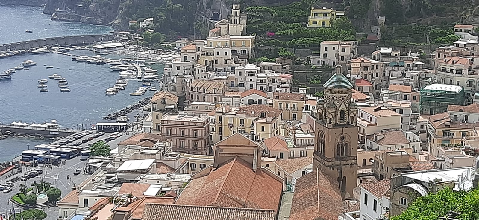 Panoramiczny widok na miasto Amalfi we Włoszech, z portem pełnym łodzi i charakterystyczną architekturą z dominującą wieżą kościelną z zieloną kopułą.
