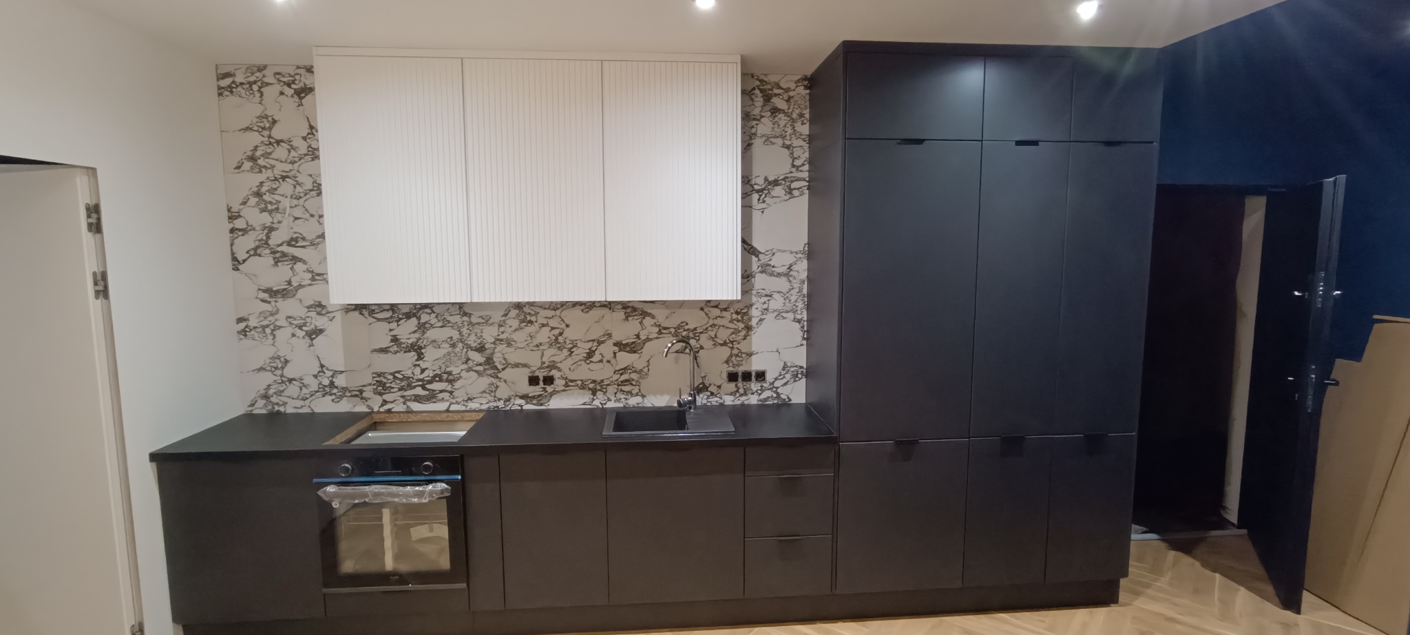 Nowoczesna kuchnia z czarnymi szafkami i blatem, białymi górnymi szafkami z ryflowanym frontem oraz marmurowym panelem ściennym. Widoczny zlew i piekarnik.