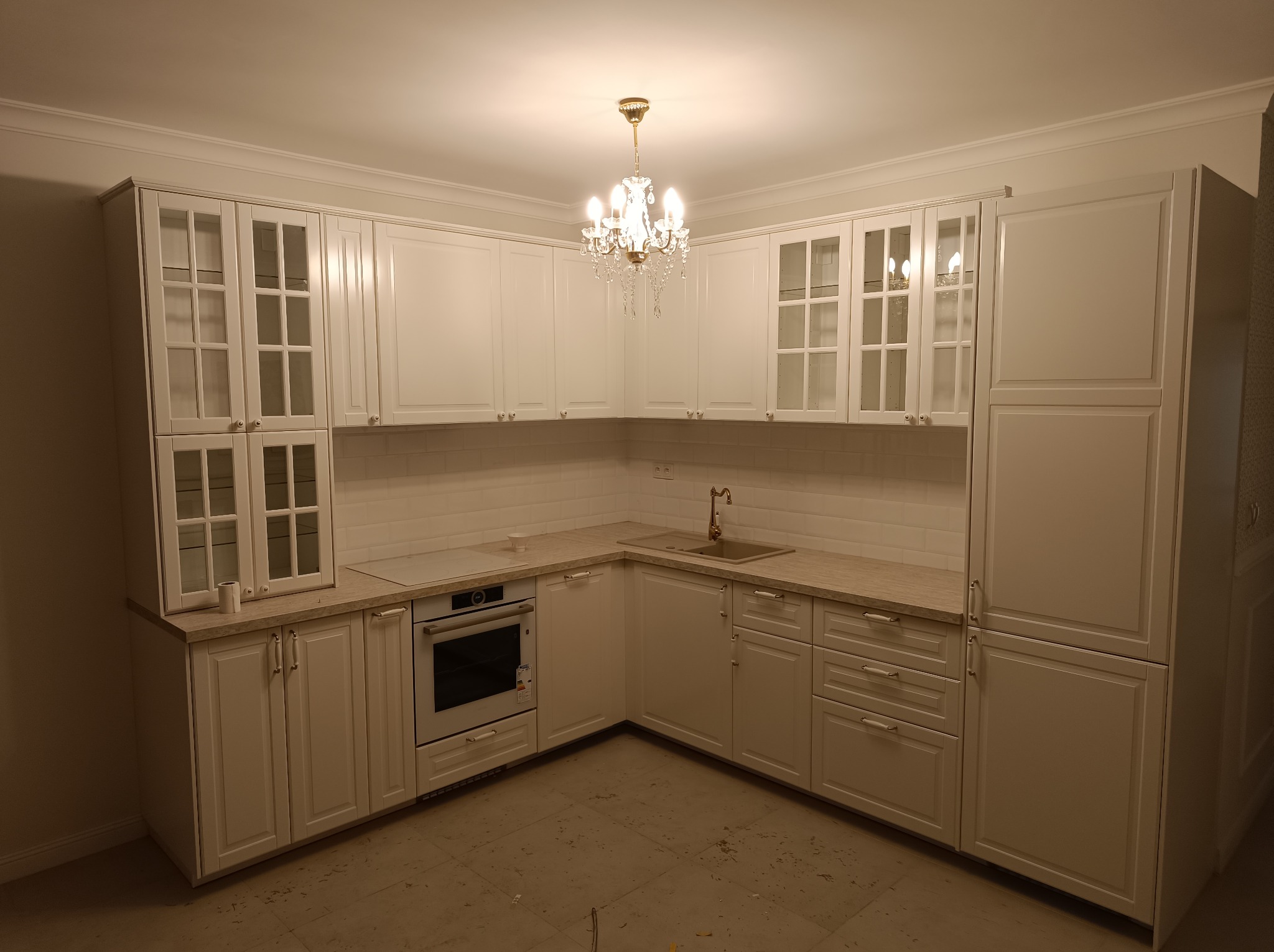 Jasna, klasyczna kuchnia z białymi szafkami, szklanymi witrynami i złotym żyrandolem. Widoczny piekarnik i zlewozmywak. Uwagę przykuwają detale wykończenia.