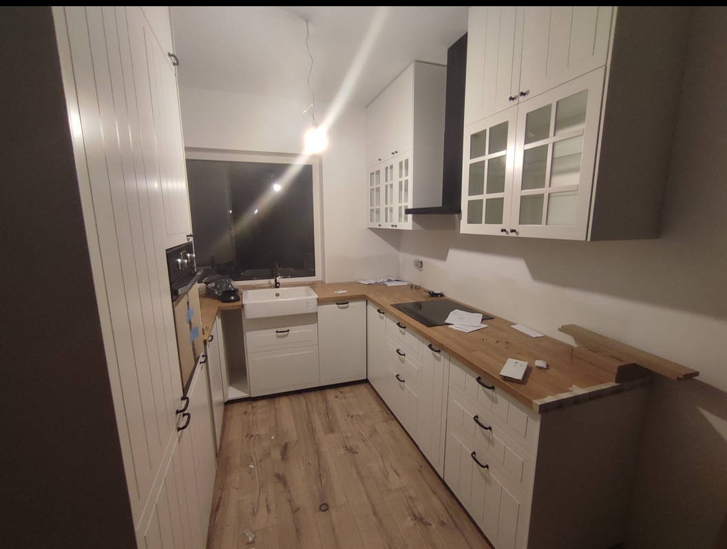 Biała kuchnia w stylu rustykalnym z drewnianym blatem, czarnym okapem i oknem. Widoczne niedokończone elementy blatu i podłogi, sugerujące etap montażu.