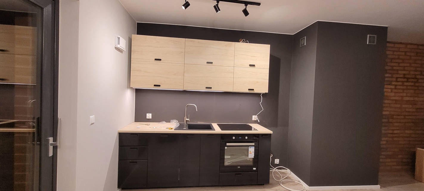 Nowoczesna kuchnia z czarnymi szafkami dolnymi i jasnymi górnymi, oświetlenie punktowe, szara ściana i fragment ściany z cegły w tle. Minimalistyczny design.