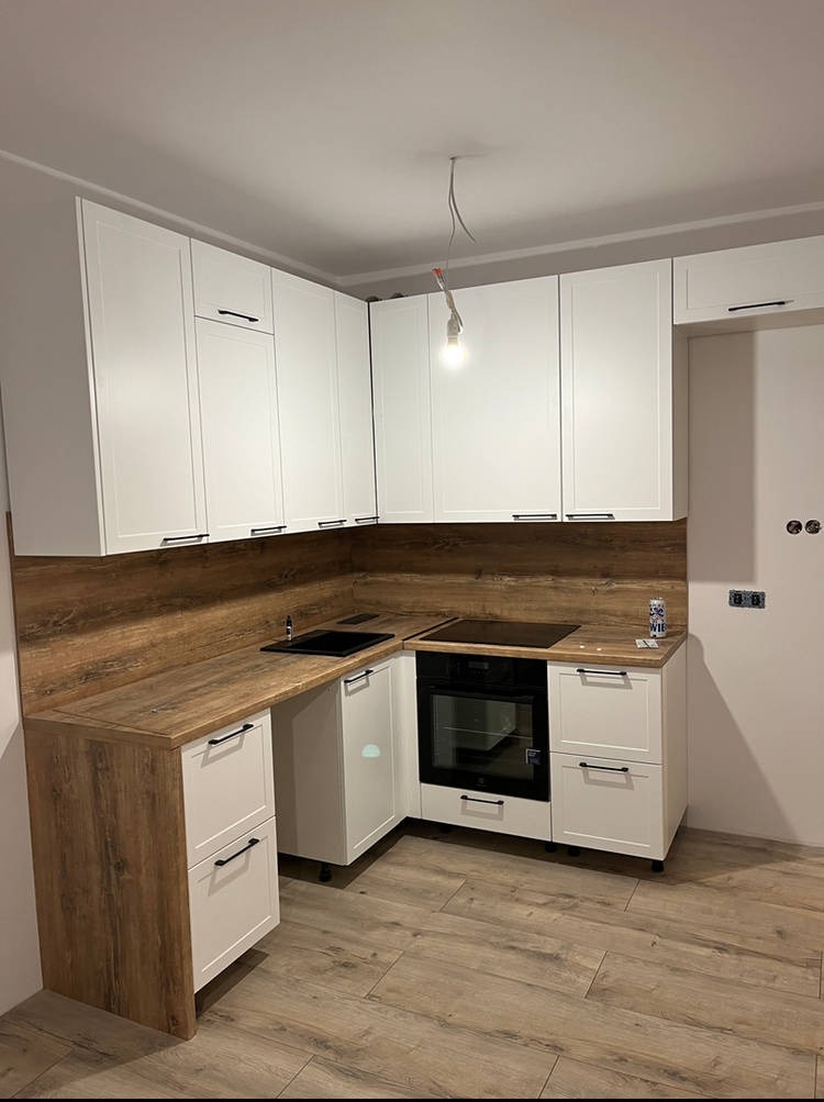 Nowoczesna kuchnia z białymi szafkami i drewnianym blatem. Widoczny zlew, płyta indukcyjna i piekarnik. Podłoga wyłożona panelami imitującymi drewno.