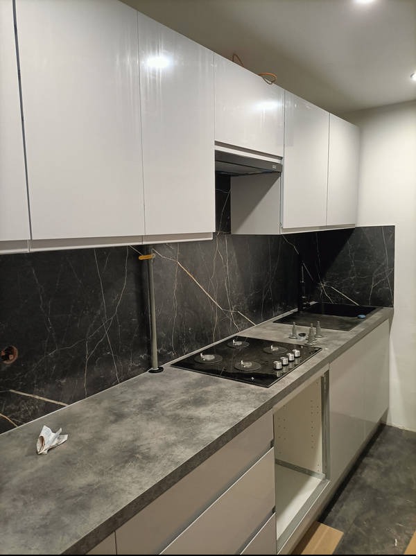 Nowoczesna kuchnia z białymi szafkami, blatem imitującym beton i czarnym panelem ściennym z marmurowym wzorem. Widoczna płyta grzewcza i zlew.