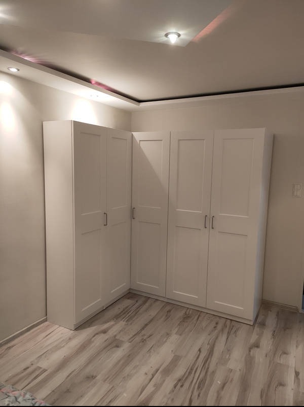 Biała, narożna szafa z czterema drzwiami, z metalowymi uchwytami, na drewnianej podłodze, w jasnym pomieszczeniu z oświetleniem sufitowym.