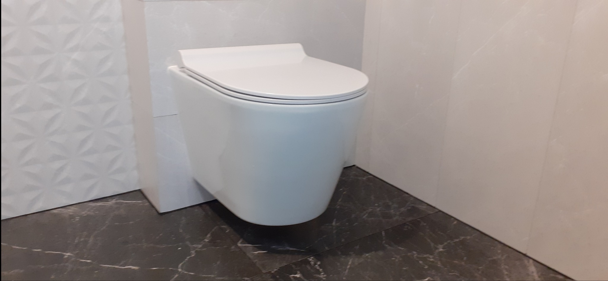 Podwieszana, biała miska WC z deską wolnoopadającą, zamontowana w nowoczesnej łazience z marmurową posadzką i ścianami wykończonymi płytkami o różnej fakturze.