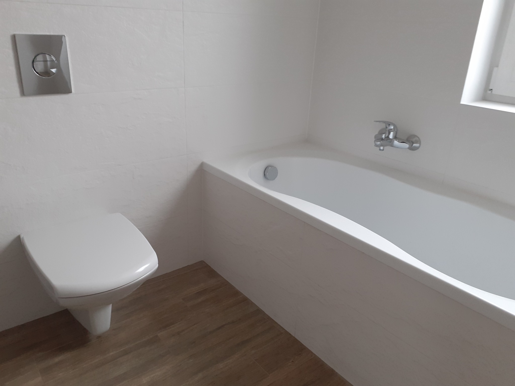 Nowoczesna, minimalistyczna łazienka z podwieszaną toaletą, białą wanną i drewnopodobną podłogą, wykończona jasnymi płytkami.