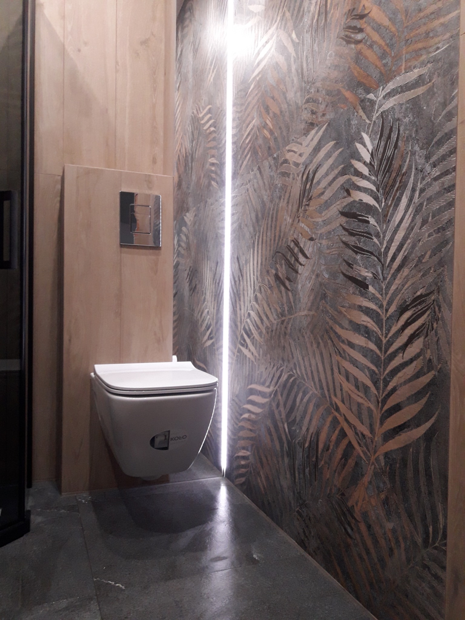 Nowoczesna łazienka z podwieszaną toaletą marki Koło, drewnopodobną zabudową, oświetleniem LED pionowym i płytkami z motywem liści palmowych.