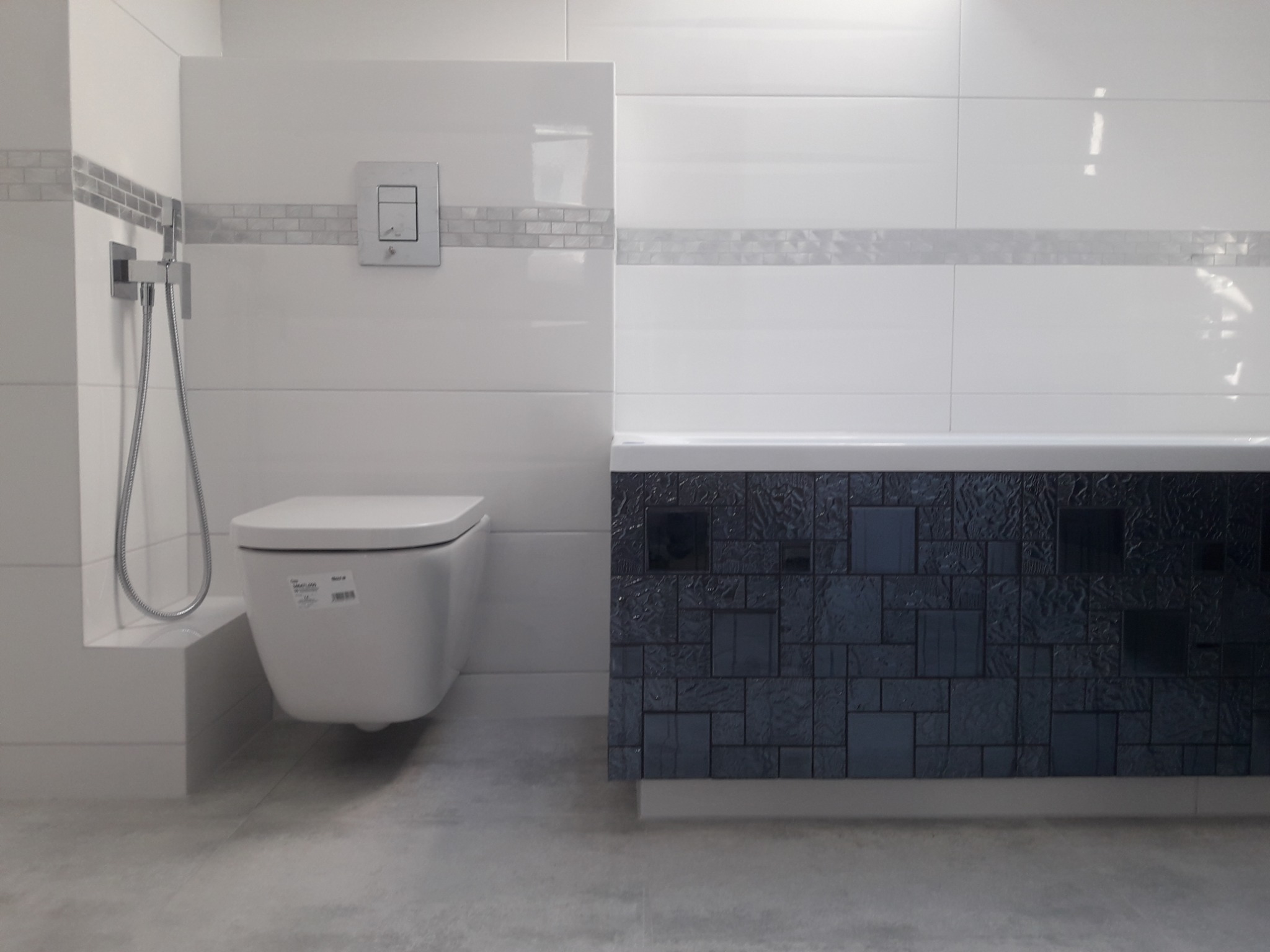 Nowoczesna łazienka z podwieszaną toaletą, bateria prysznicowa z wężem na białej ścianie, dekoracyjna mozaika w pasach, oraz obudowa wanny z ciemnych płytek o zróżnicowanej fakturze.