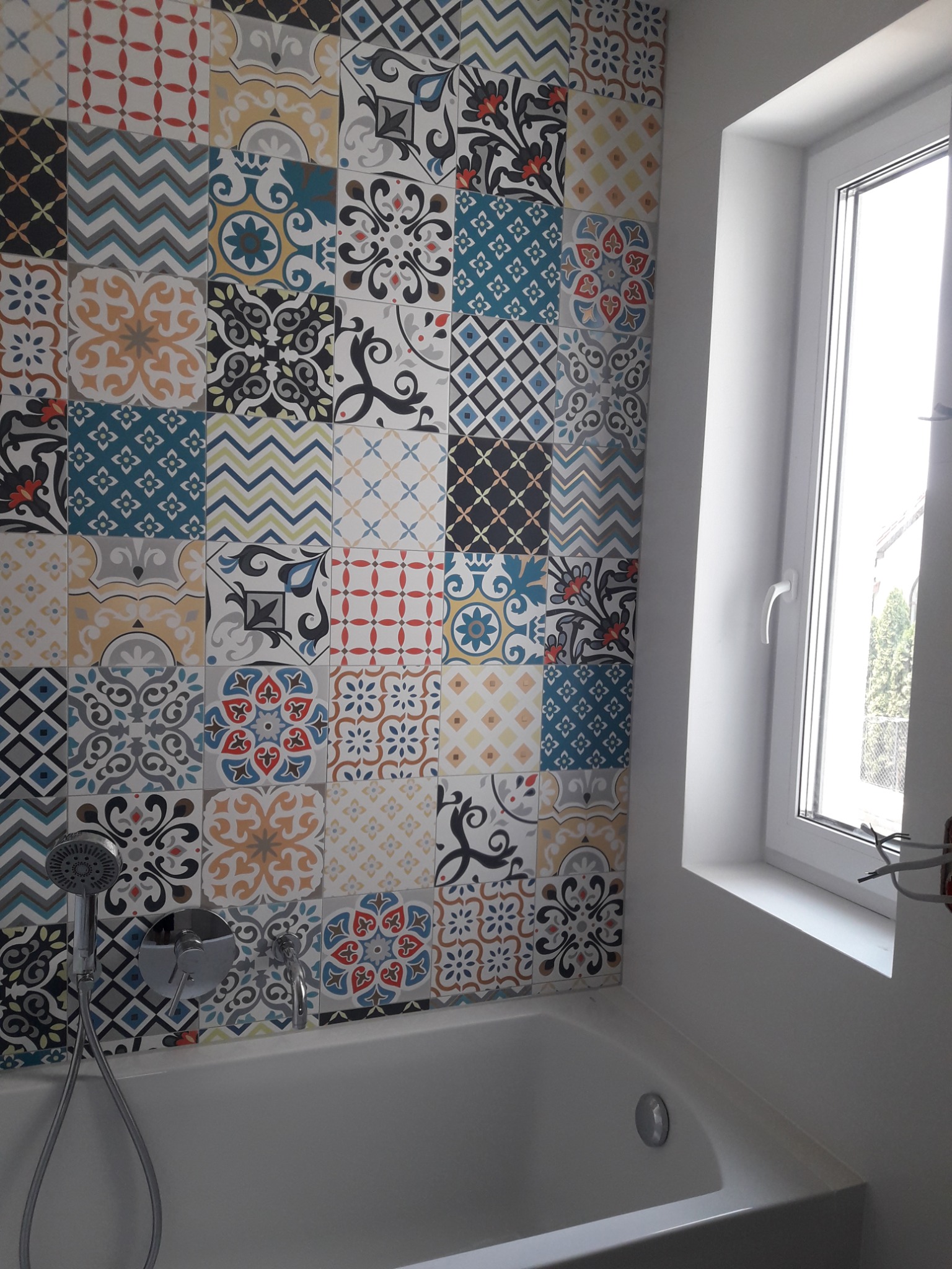 Wanna z baterią i słuchawką prysznicową na tle ściany wyłożonej wielobarwnymi, wzorzystymi płytkami ceramicznymi; obok okno z białymi ramami.