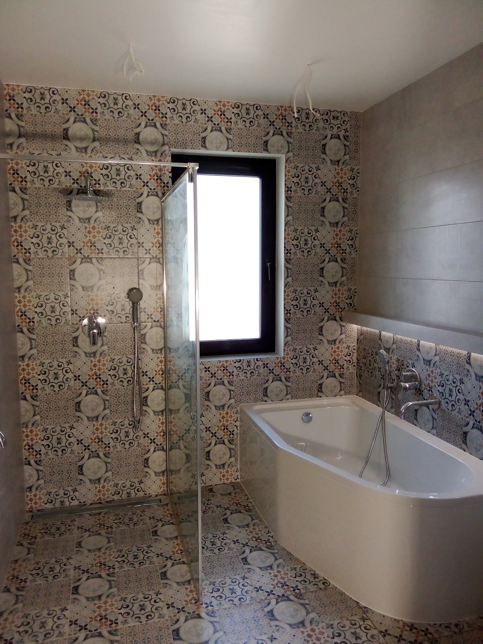 Wykończona łazienka z kabiną prysznicową typu walk-in, białą wanną narożną i wzorzystymi płytkami na ścianach i podłodze, widoczne wystające kable z sufitu.