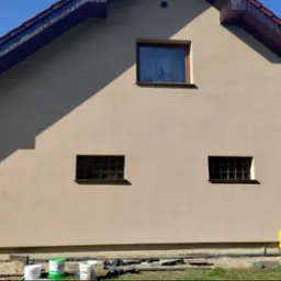 Fragment domu z beżową elewacją, widoczne okna, skrzynka gazowa i puste wiadra po farbie na trawie, cień rzucany przez dach.