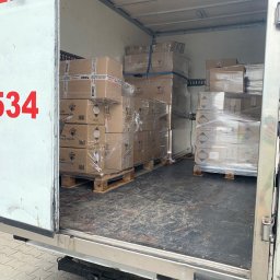 Easy Movers - Wnętrze busa transportowego załadowane paletami z kartonami, zabezpieczonymi folią stretch i pasami, widoczne etykiety ostrzegawcze i nazwy produktów (Mr. Camp, Profast, Chemia Basenowa).