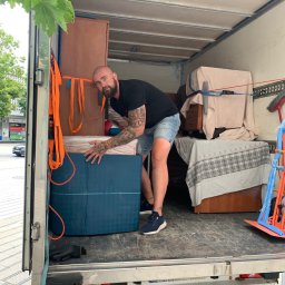 Easy Movers - Mężczyzna z tatuażami wewnątrz furgonetki ładuje zapakowane meble, zabezpieczone pasami transportowymi, widoczne kartony i fragmenty szaf.