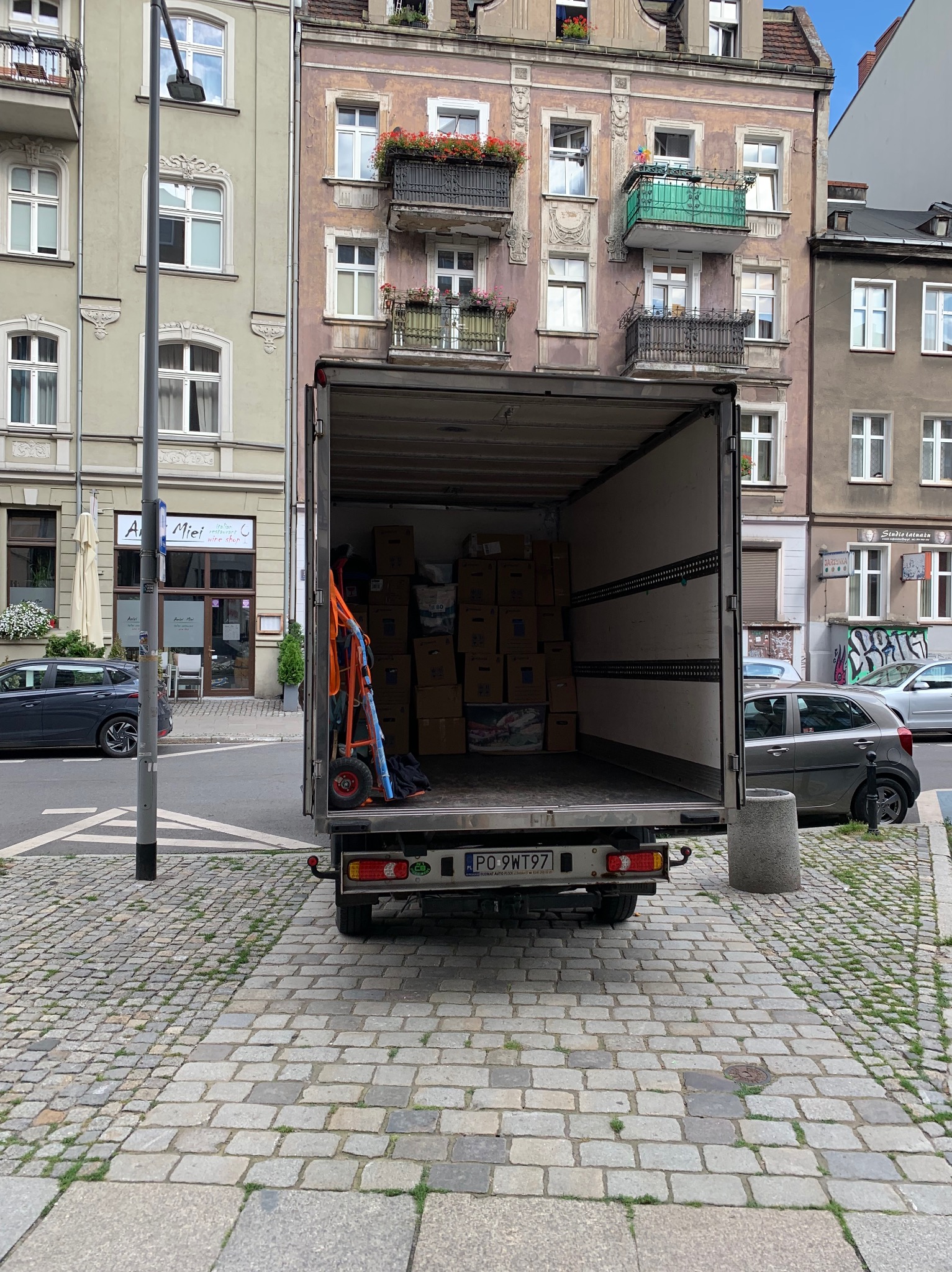 Otwarta paka dostawczego busa załadowana kartonami, w tle kamienica z balkonami i kwiatami, widoczny wózek transportowy z pomarańczowymi pasami.