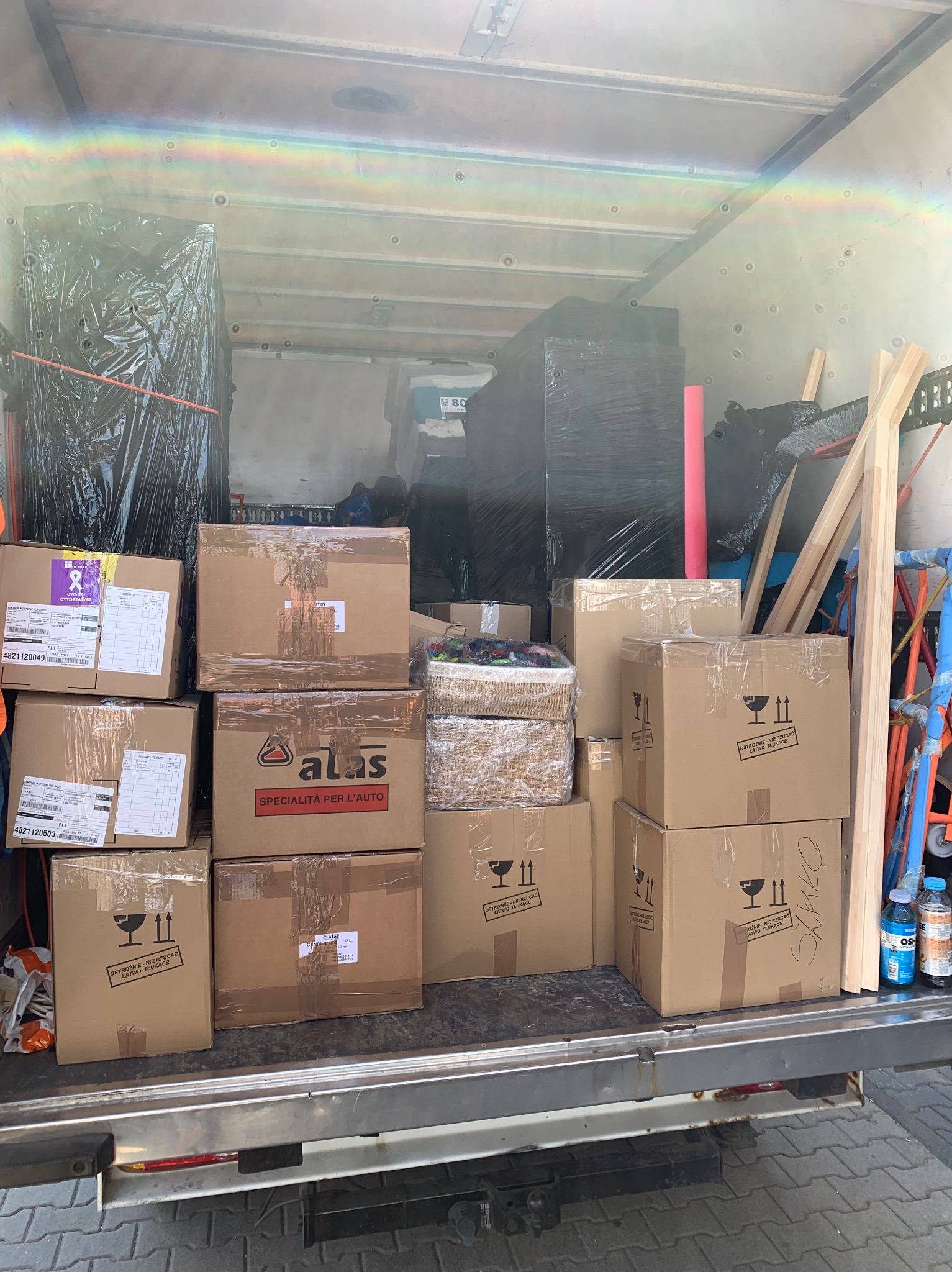 Wnętrze wypełnionej skrzyniami ładowni busa transportowego; zabezpieczone meble w folii stretch na tle, z przodu kartony z oznaczeniami ostrożności i adresami.