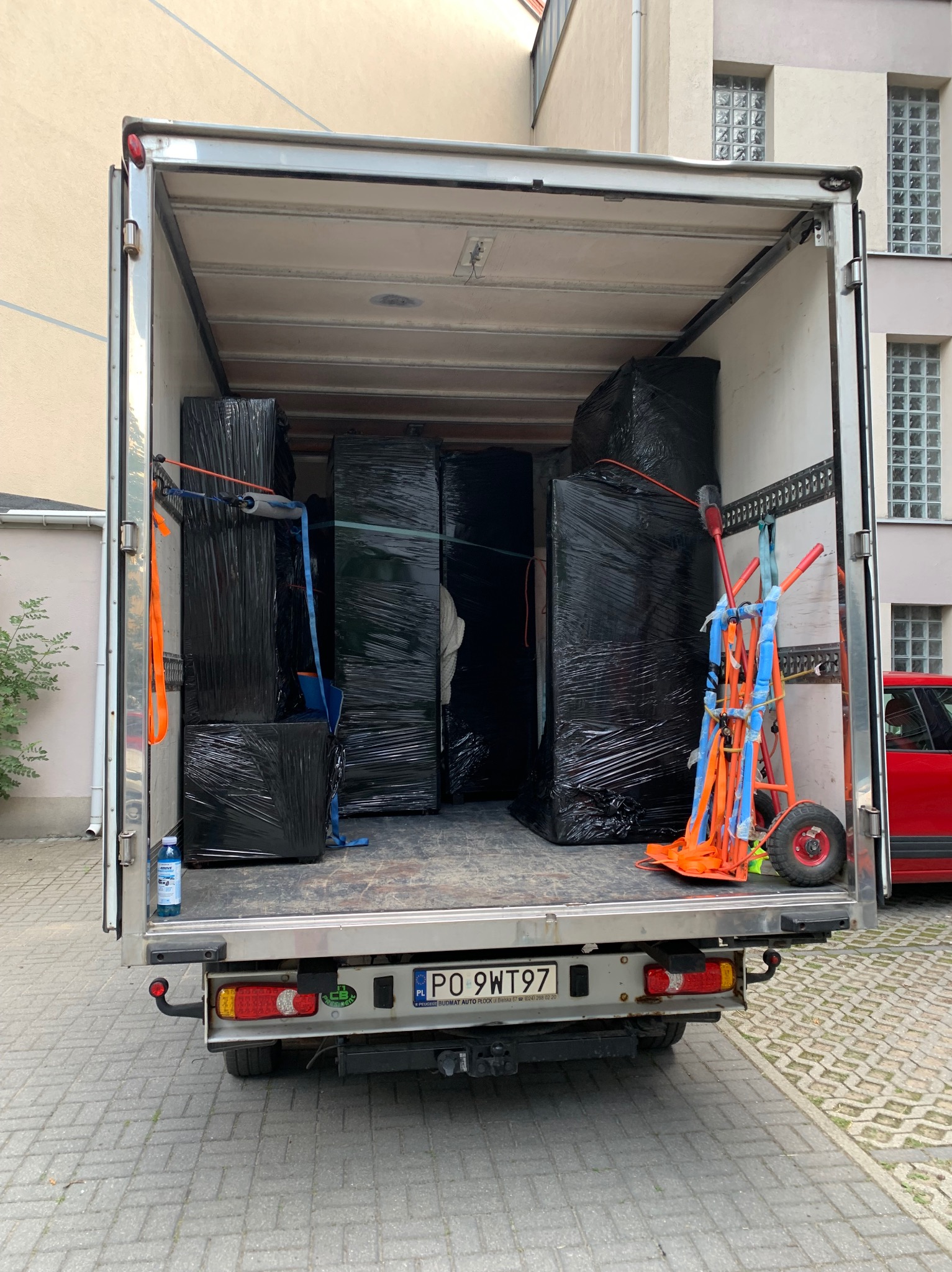 Wnętrze busa transportowego załadowane meblami owiniętymi czarną folią stretch, zabezpieczonymi pasami, w tle wózek transportowy, widok z tyłu pojazdu z otwartymi drzwiami.