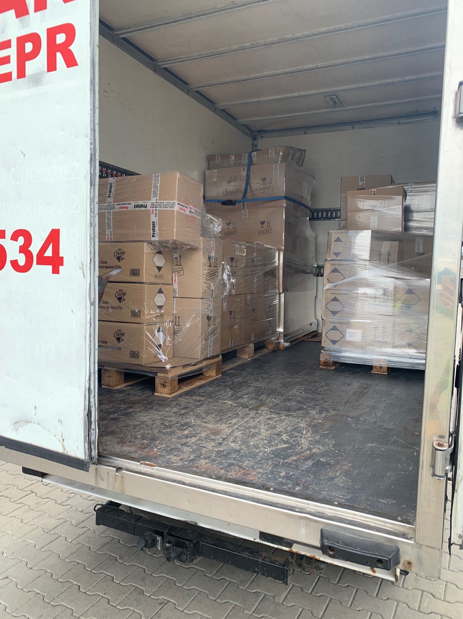 Wnętrze busa transportowego załadowane paletami z kartonami, zabezpieczonymi folią stretch i pasami, widoczne etykiety ostrzegawcze i nazwy produktów (Mr. Camp, Profast, Chemia Basenowa).