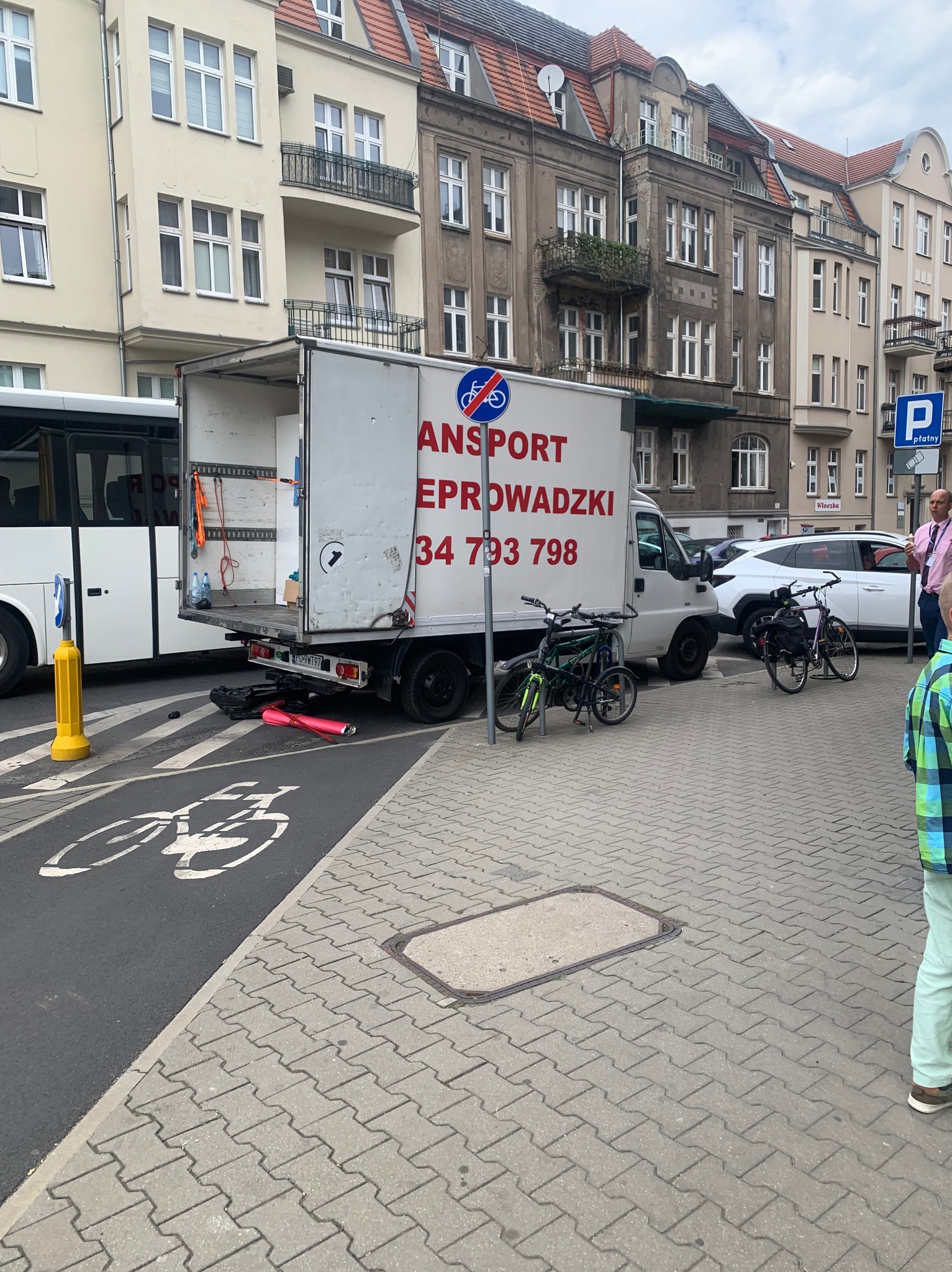 Otwarty tył białej furgonetki z napisem 'ANSPORT EPROWADZKI' zaparkowanej na ulicy miasta, obok ścieżki rowerowej i zaparkowanych rowerów.