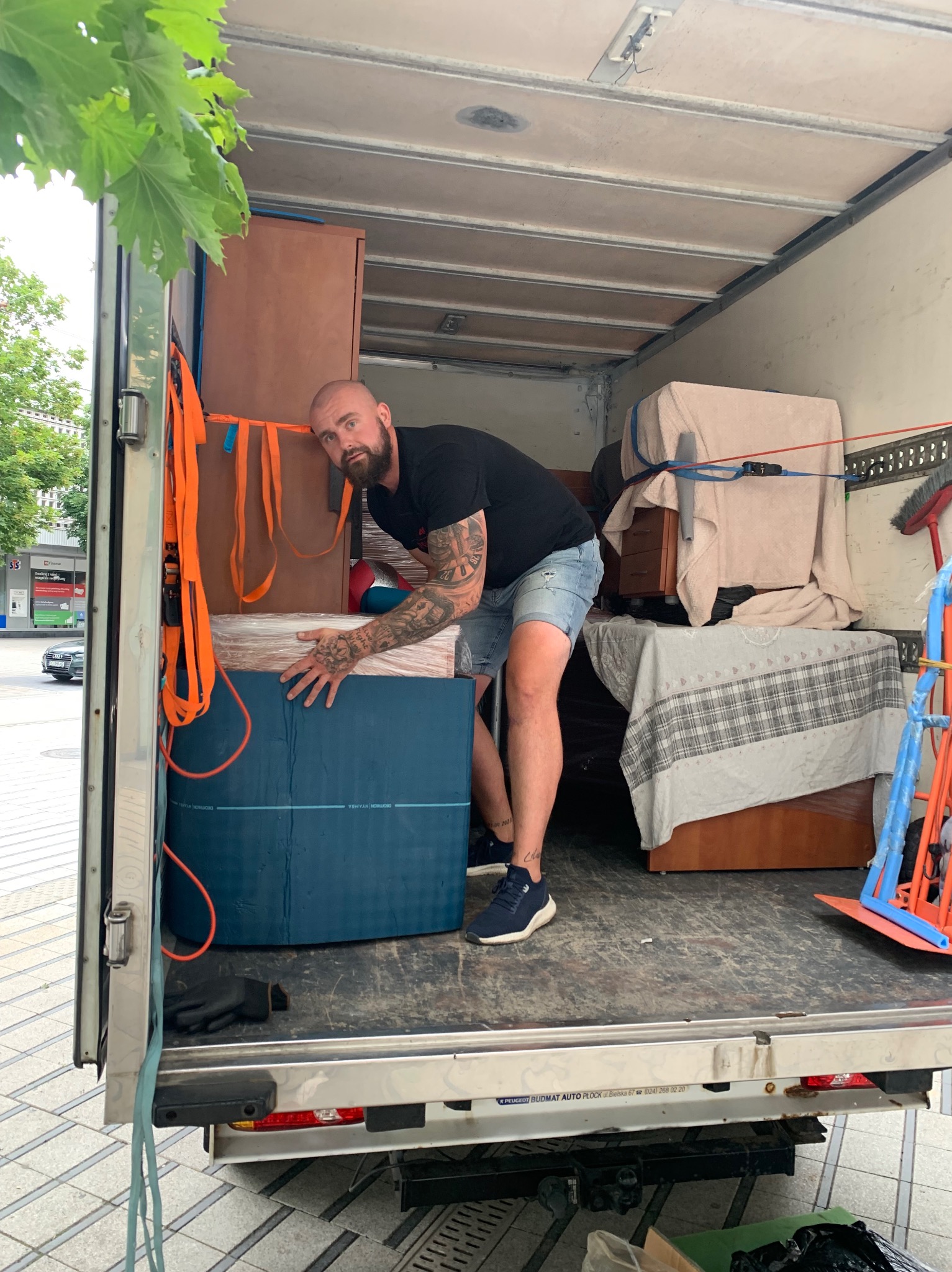 Mężczyzna z tatuażami wewnątrz furgonetki ładuje zapakowane meble, zabezpieczone pasami transportowymi, widoczne kartony i fragmenty szaf.