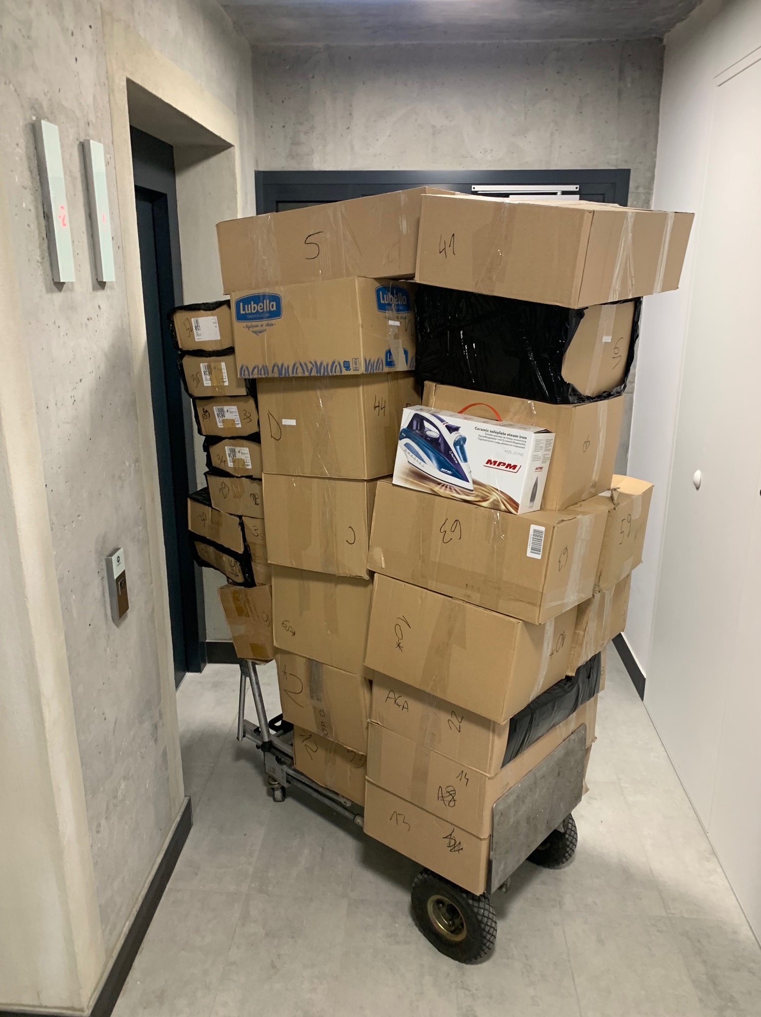 Wózek transportowy obładowany kartonami w korytarzu budynku mieszkalnego, widoczna również paczka z żelazkiem parowym MPM.