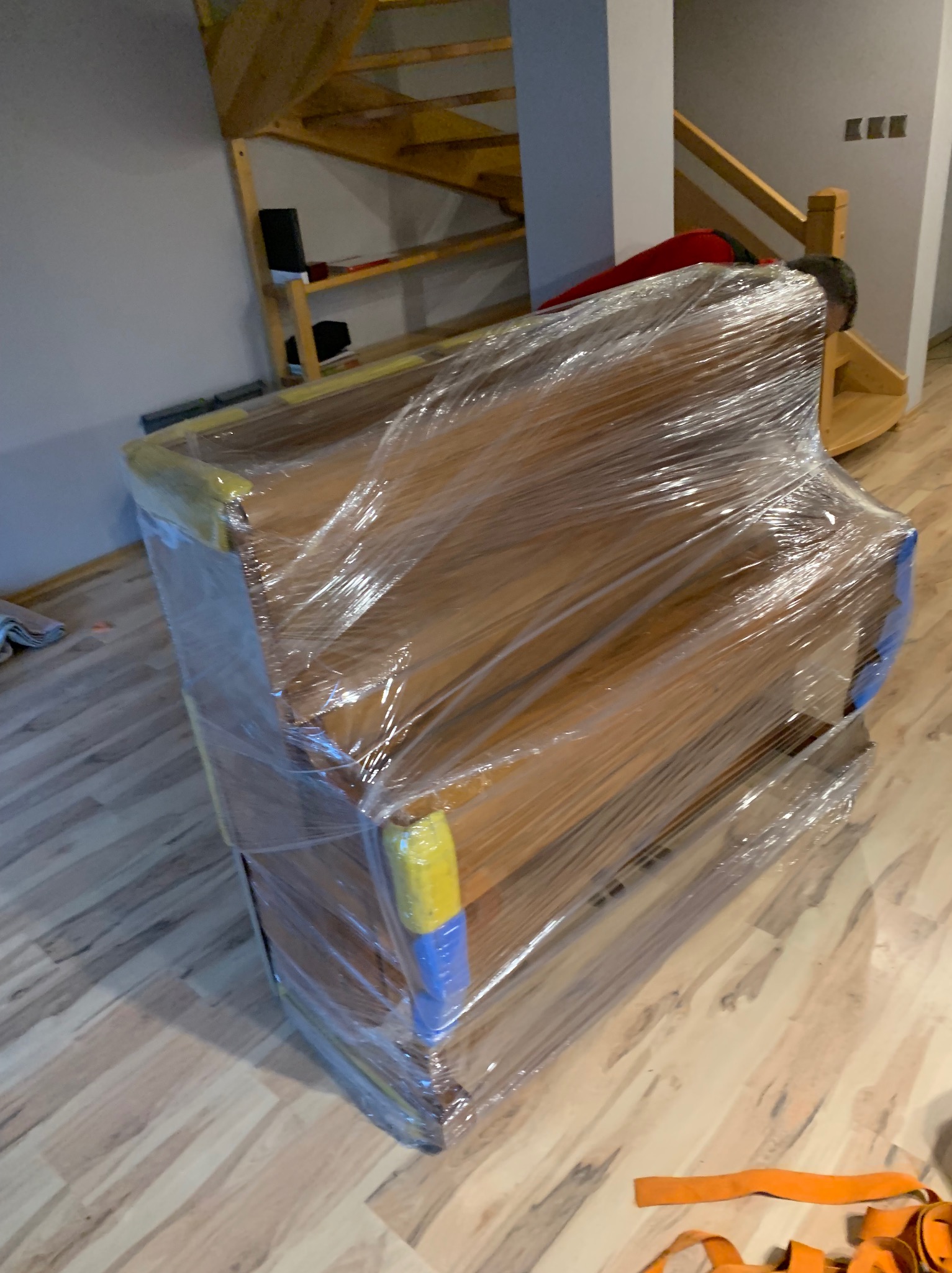 Pianino zabezpieczone folią stretch i taśmą w kolorach żółtym i niebieskim, przygotowane do transportu wewnątrz domu z drewnianymi schodami.