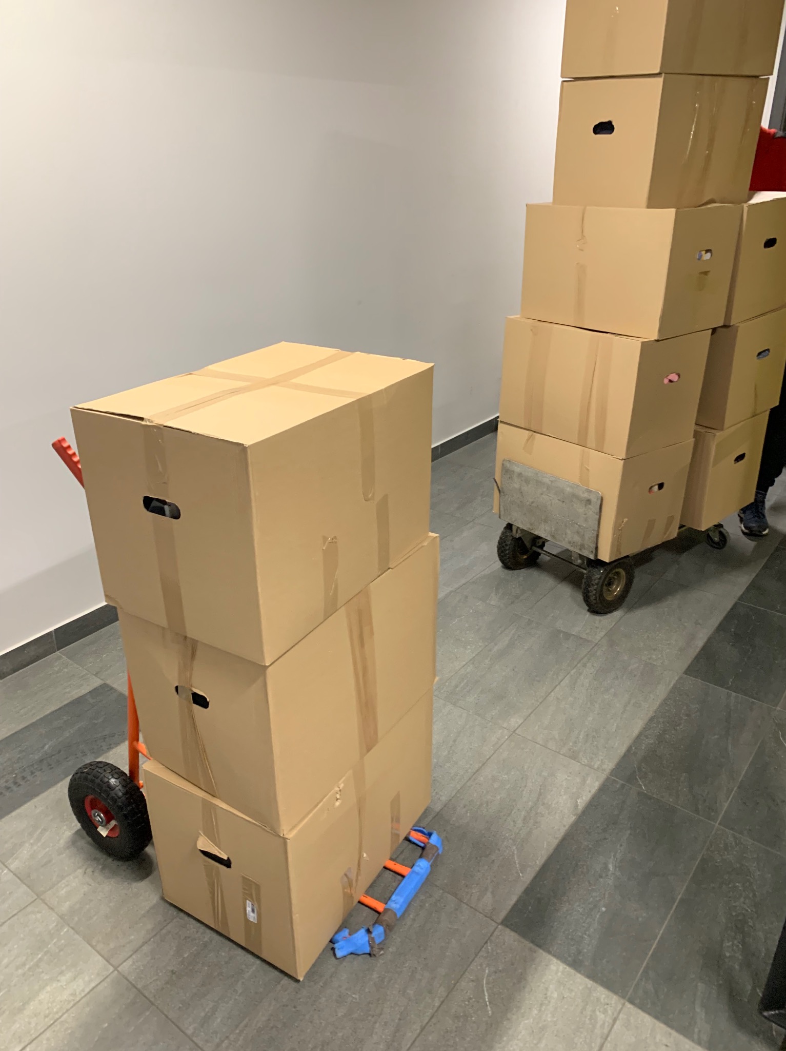 Stos kartonowych pudeł z uchwytami, zabezpieczonych taśmą, na wózku transportowym, przygotowanych do przeprowadzki, z fragmentem niebieskiego wózka obok