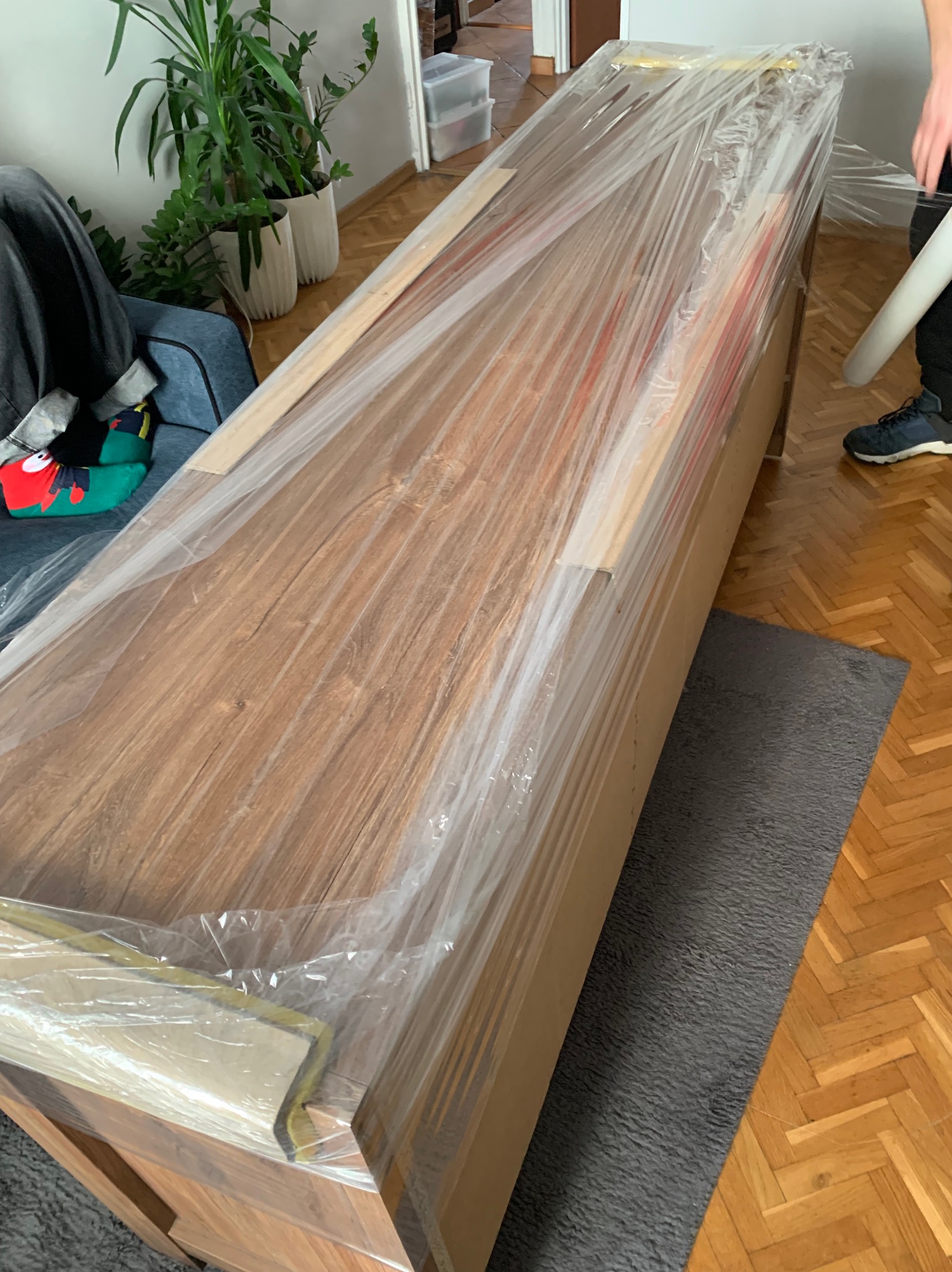 Masywny drewniany mebel, zabezpieczony folią stretch i kartonem, przygotowany do transportu w mieszkaniu z parkietem w jodełkę.