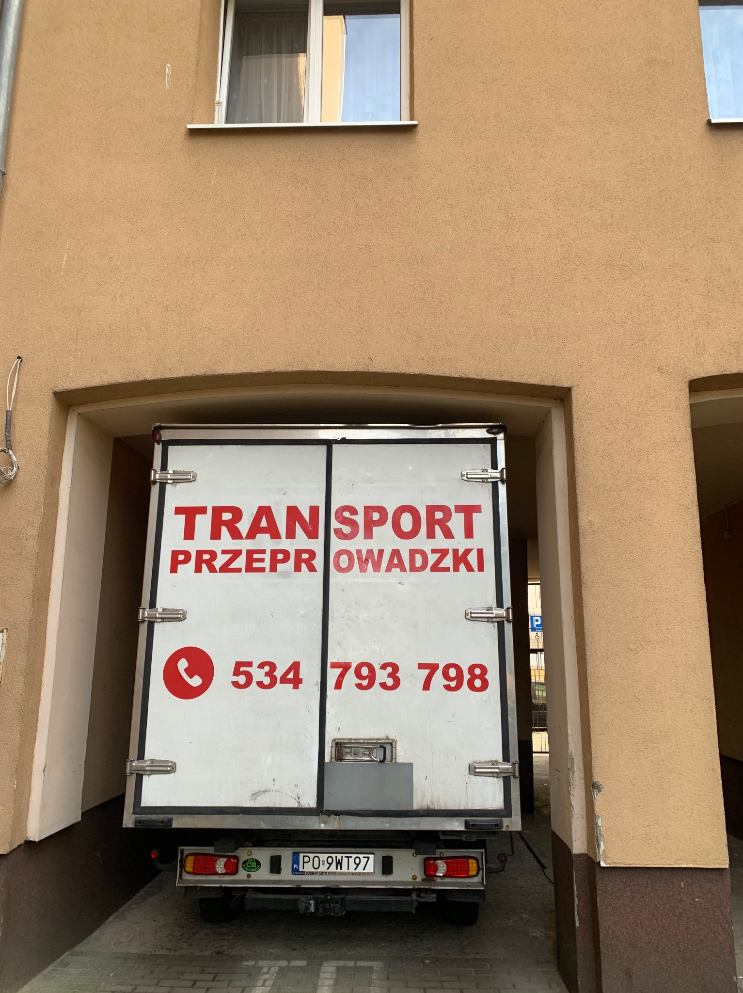 Tył białego samochodu dostawczego z czerwonym napisem 'TRANSPORT PRZEPROWADZKI' i numerem telefonu, zaparkowanego w przejeździe budynku o beżowej elewacji.