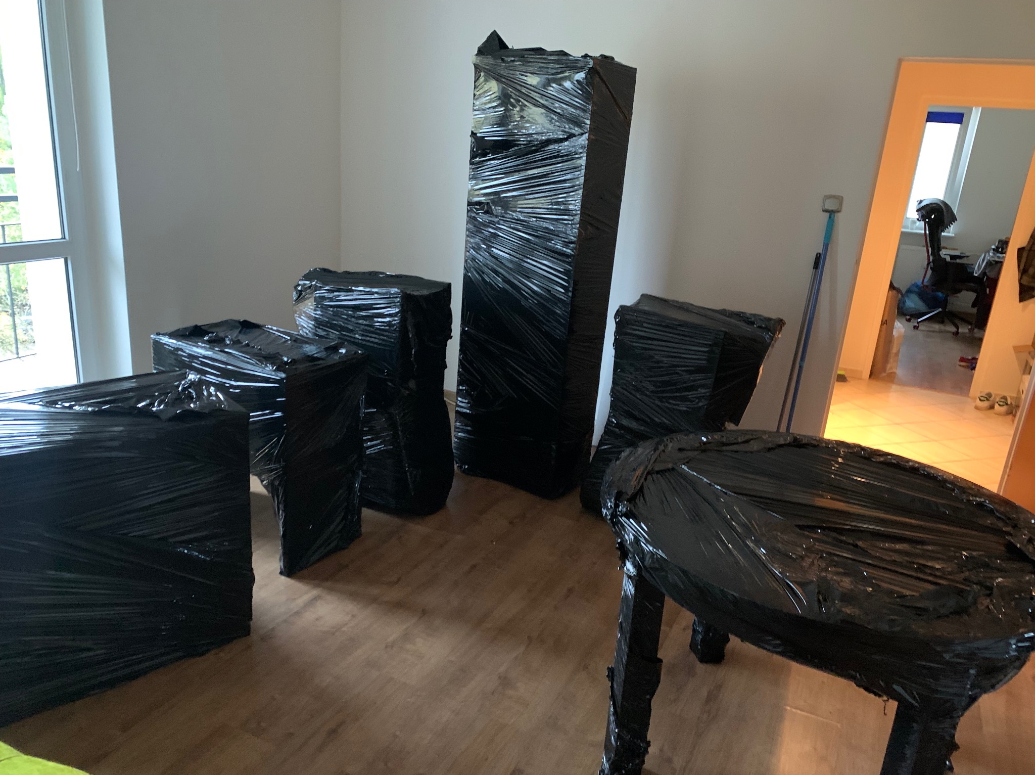 Meble owinięte czarną folią stretch, przygotowane do transportu, ustawione w pustym, jasnym pokoju z parkietem.