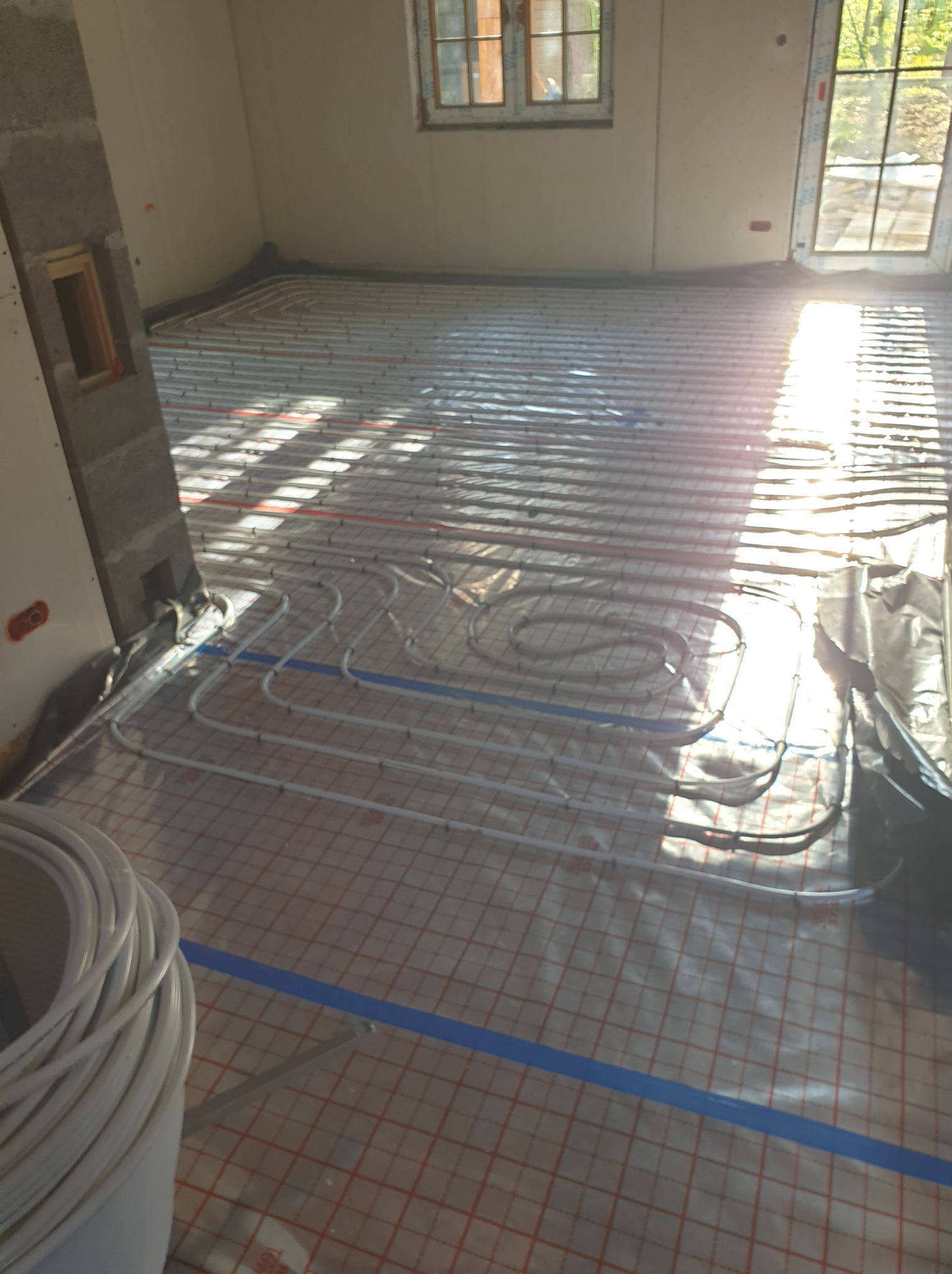 Białe rury ogrzewania podłogowego ułożone na folii z czerwoną kratką w jasnym pomieszczeniu w trakcie instalacji, widoczne światło słoneczne wpadające przez okno.
