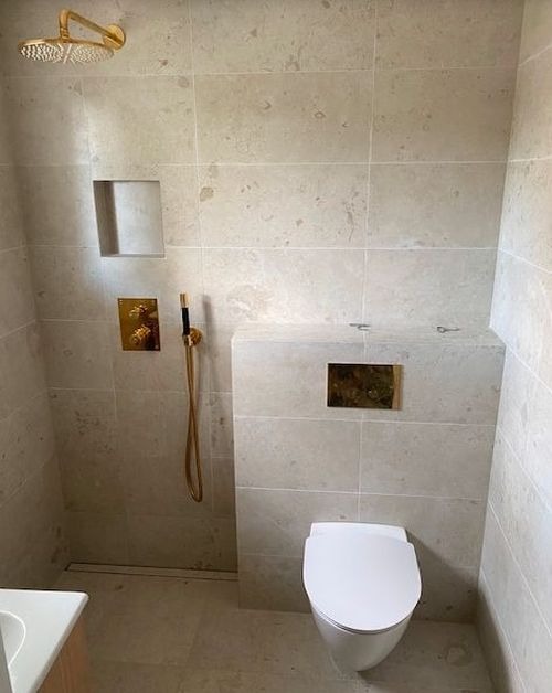 Nowoczesna łazienka wykończona płytkami imitującymi kamień, z złotą armaturą: deszczownica, bateria prysznicowa, panel sterujący; wisząca toaleta z ukrytą spłuczką i złotym przyciskiem.