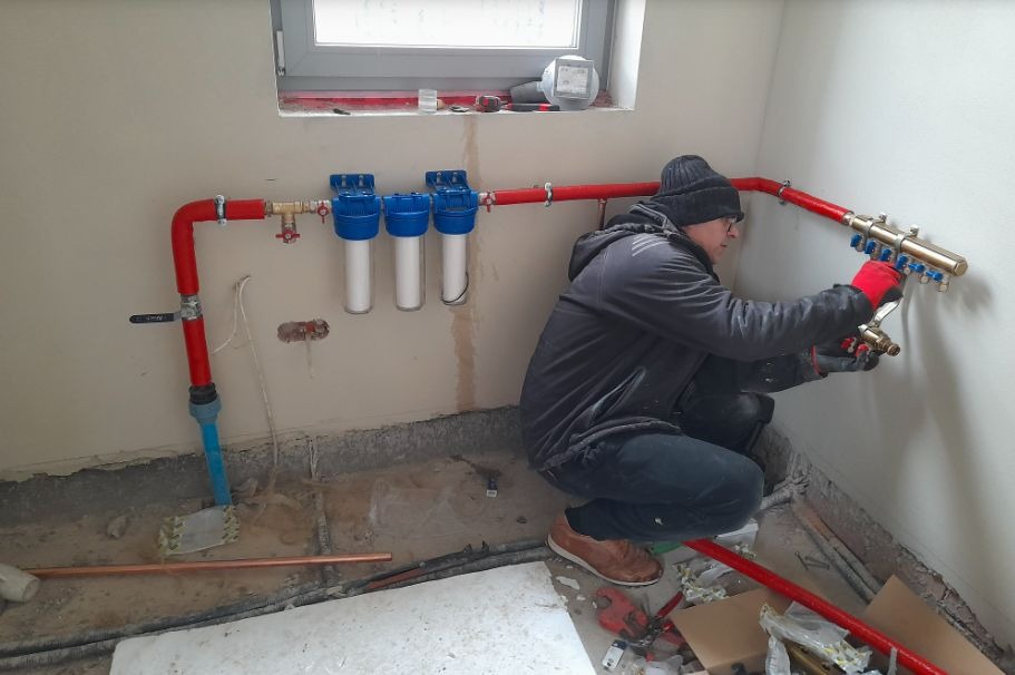Instalator w czapce i czerwonych rękawicach łączy mosiężny rozdzielacz do czerwonych rur w surowym wnętrzu z trzema filtrami wody.