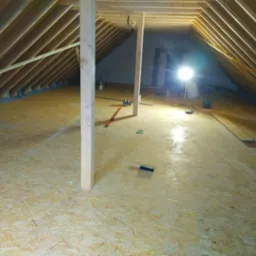 Poddasze w trakcie adaptacji: widoczne krokwie, ocieplenie między nimi, podłoga z płyt OSB, tymczasowe podpory drewniane, lampa budowlana oświetlająca przestrzeń.