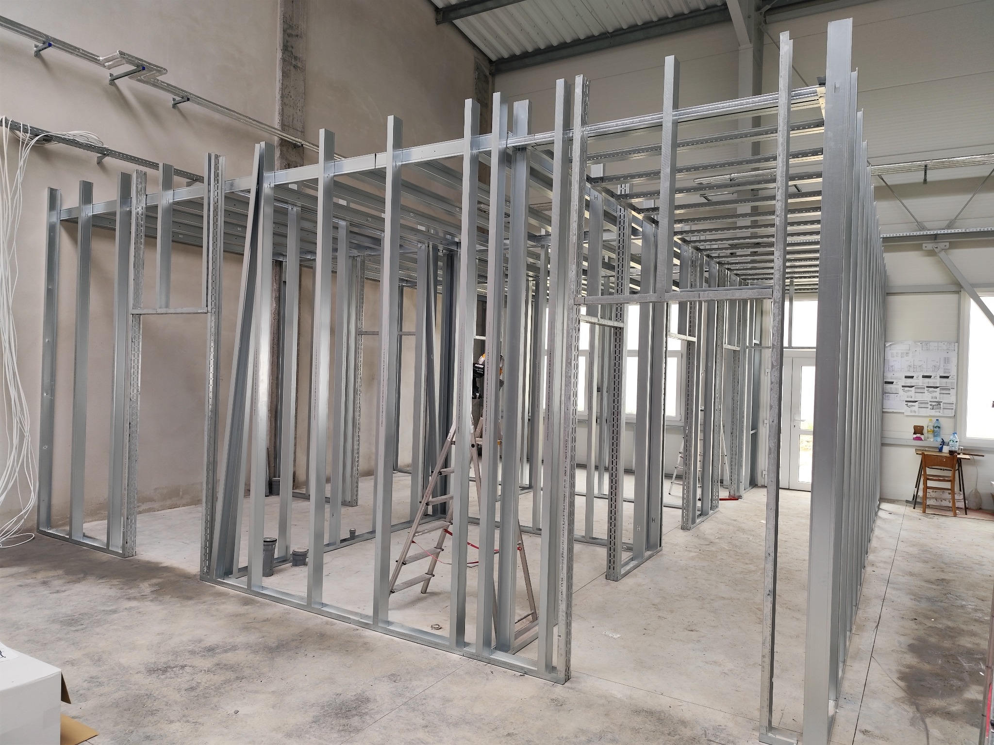 Metalowa konstrukcja szkieletowa ścian działowych w surowym stanie, widoczna drabina i instalacje pod sufitem.