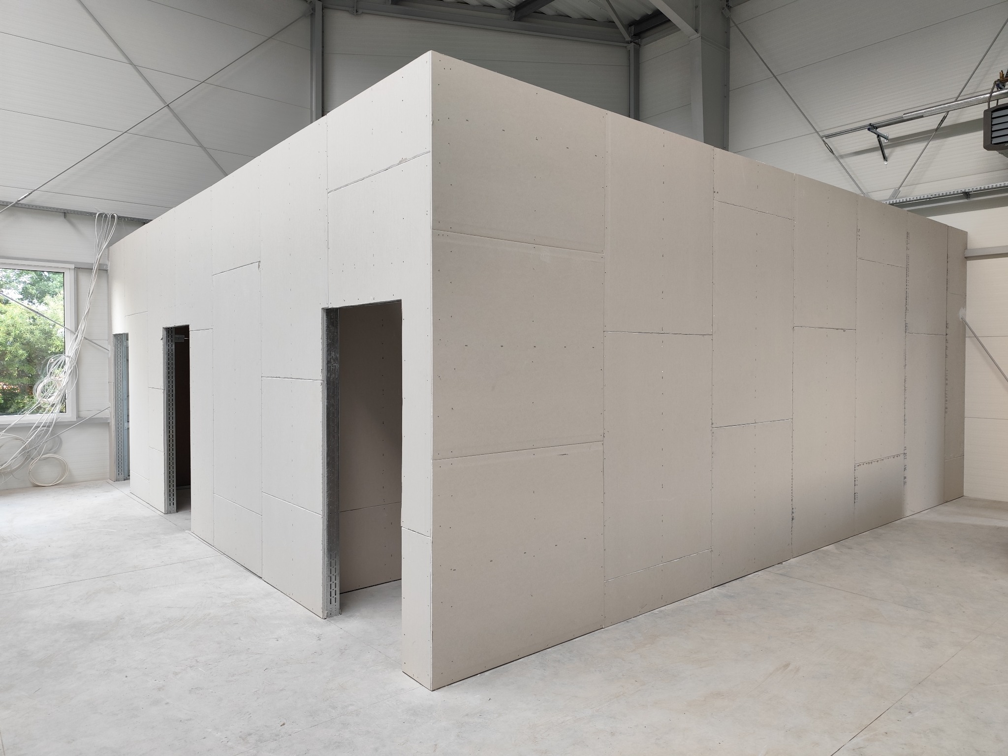 Wnętrze magazynu z wydzielonymi pomieszczeniami z płyt gipsowo-kartonowych, widoczne metalowe profile ościeżnic drzwiowych i surowa betonowa posadzka.