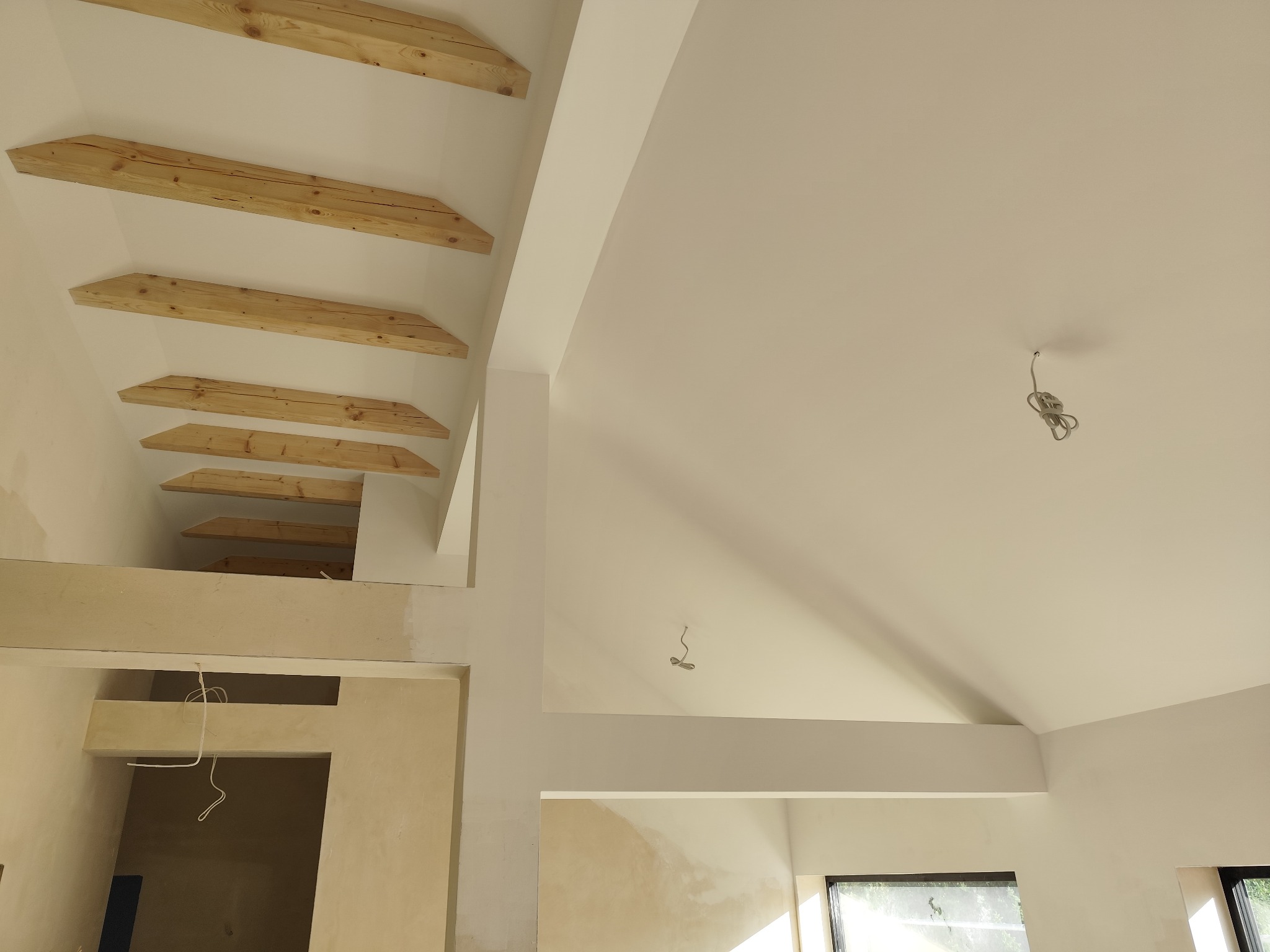 Surowe drewniane belki stropowe w jasnym wnętrzu, widoczne przewody elektryczne, ściany przygotowane do malowania.