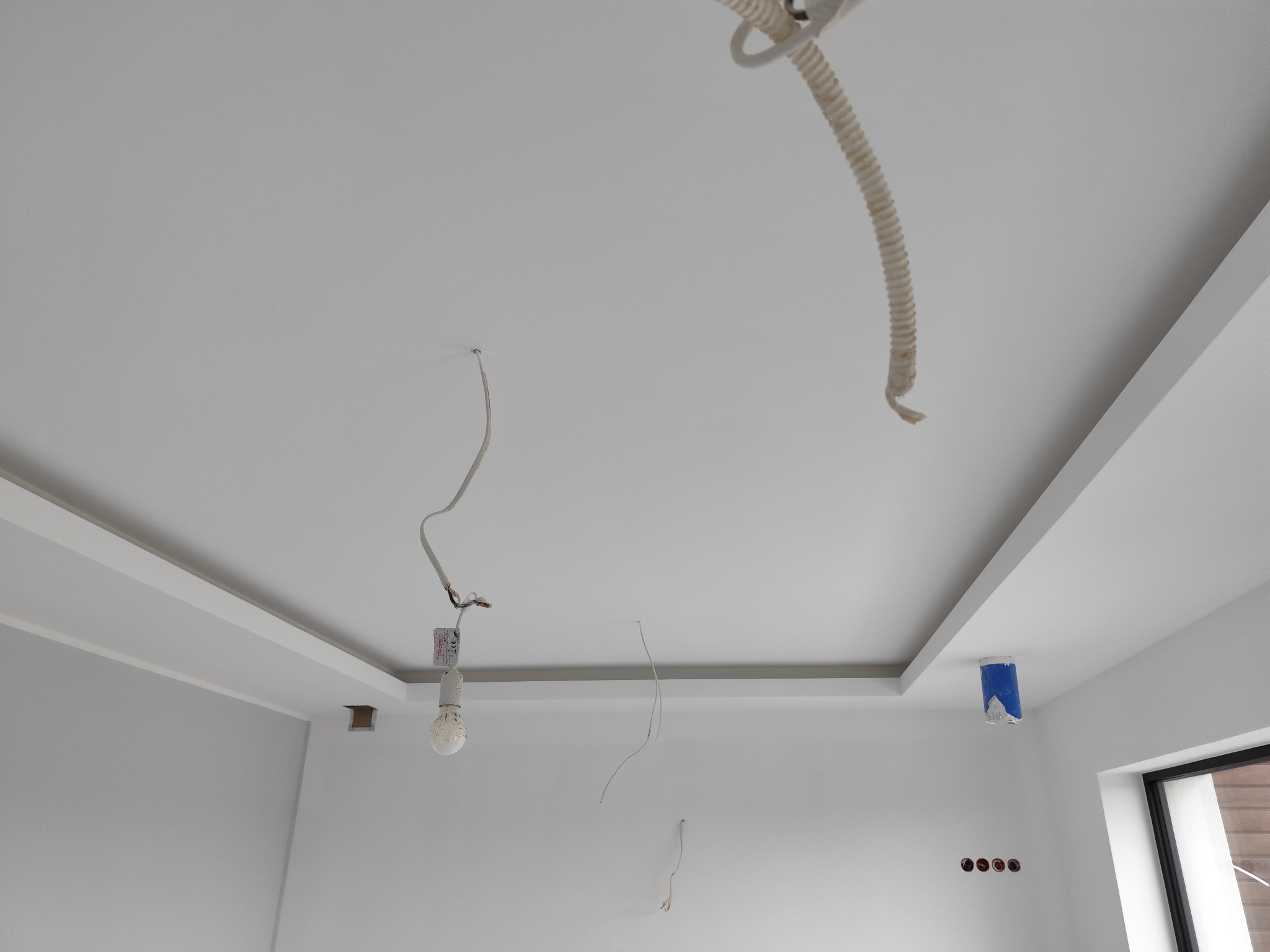 Biały sufit podwieszany z kasetonami, widoczne przewody elektryczne, prowizoryczna żarówka i zabezpieczone kable, ściany pomalowane na biało, okno z czarną ramą.