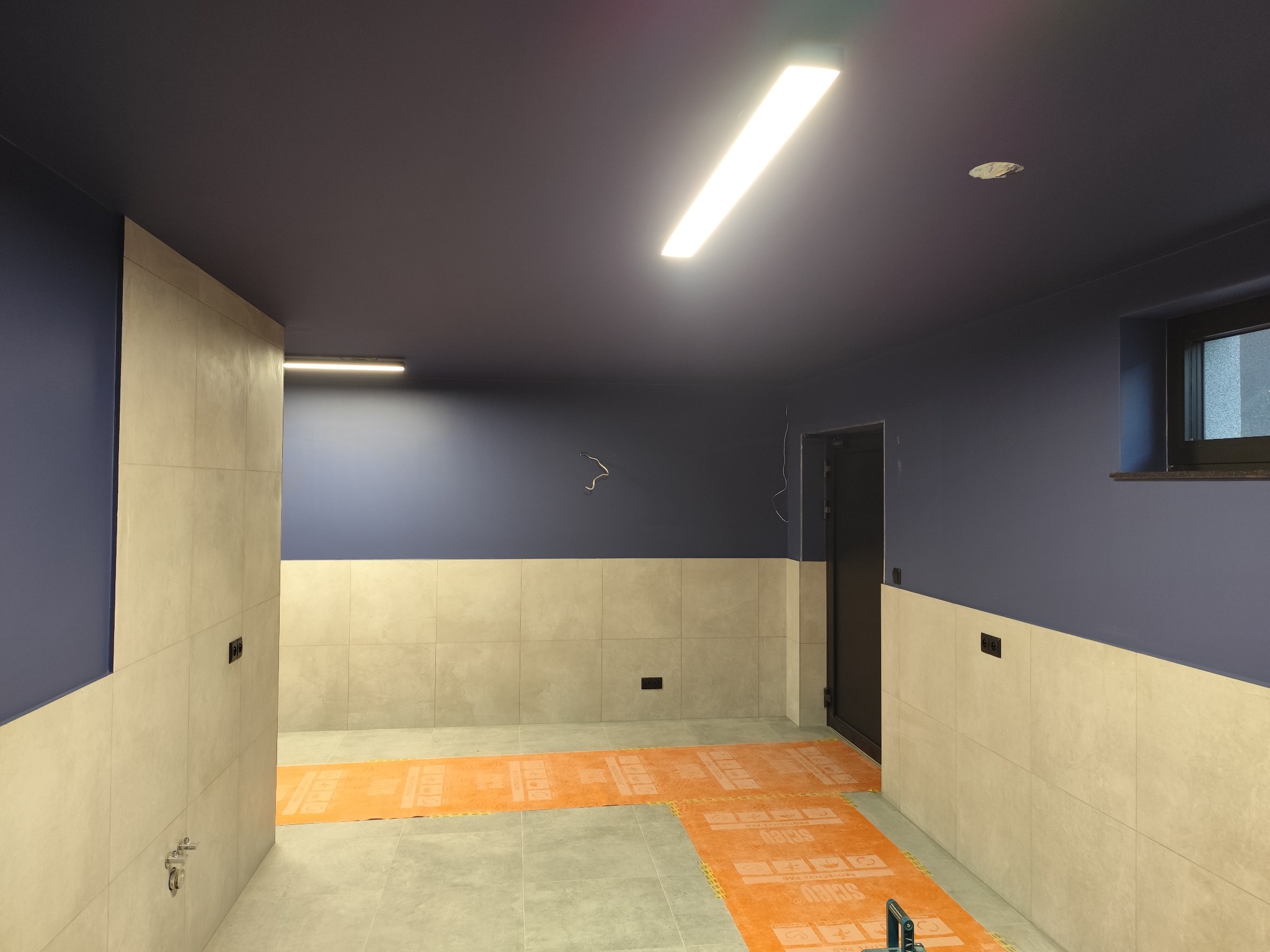 Wnętrze w trakcie prac wykończeniowych: ciemny sufit, jasne płytki na ścianach do połowy wysokości oraz pomarańczowa mata ochronna na podłodze.