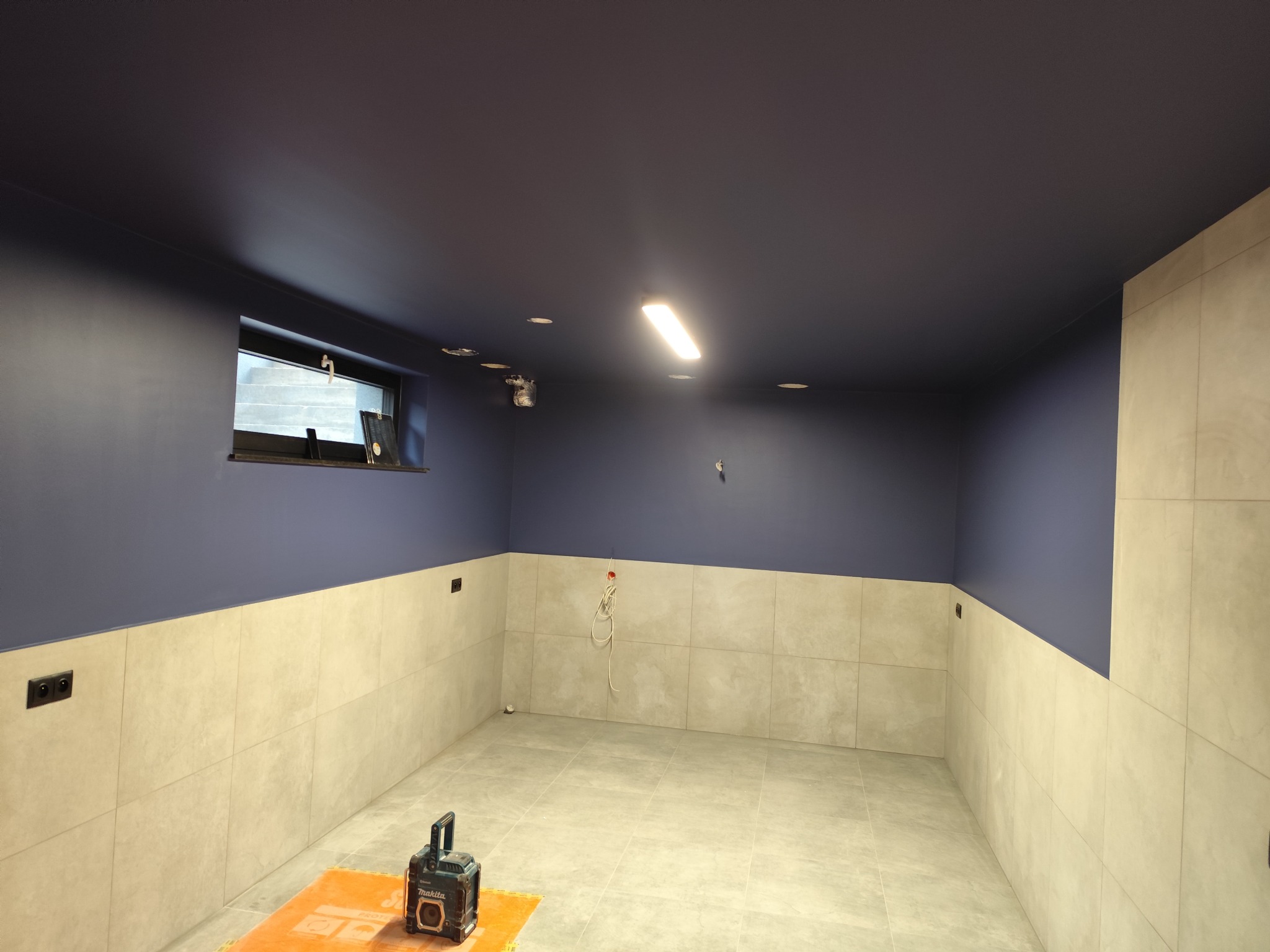 Wykończone wnętrze piwnicy z ciemnoniebieskim sufitem, jasnymi płytkami na ścianach i podłodze. Widoczne gniazdka elektryczne i narzędzie na podłodze.
