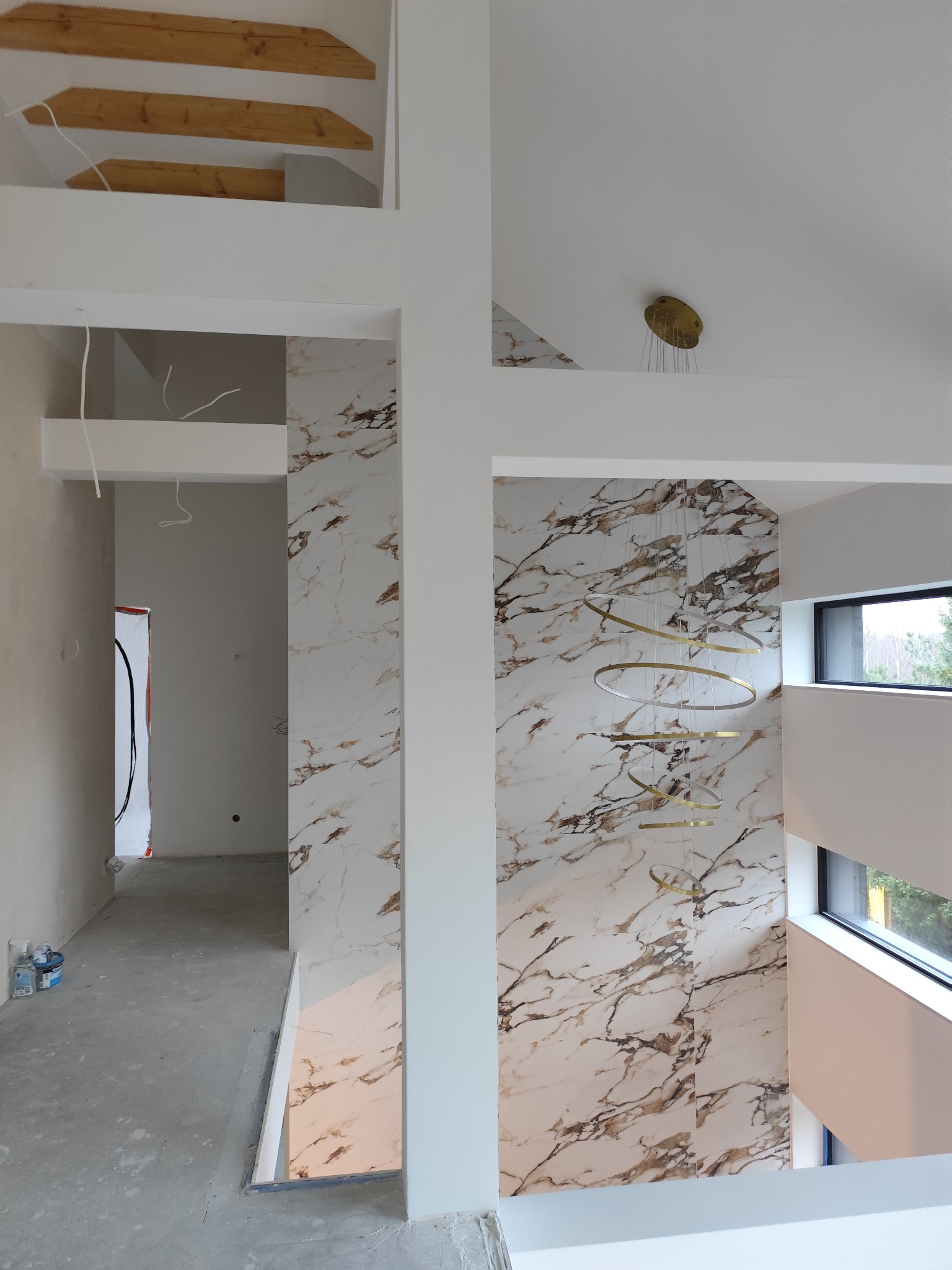 Wnętrze w trakcie wykańczania: marmurowe ściany, surowy beton na podłodze, nowoczesna lampa wisząca i drewniane elementy konstrukcyjne na górze.