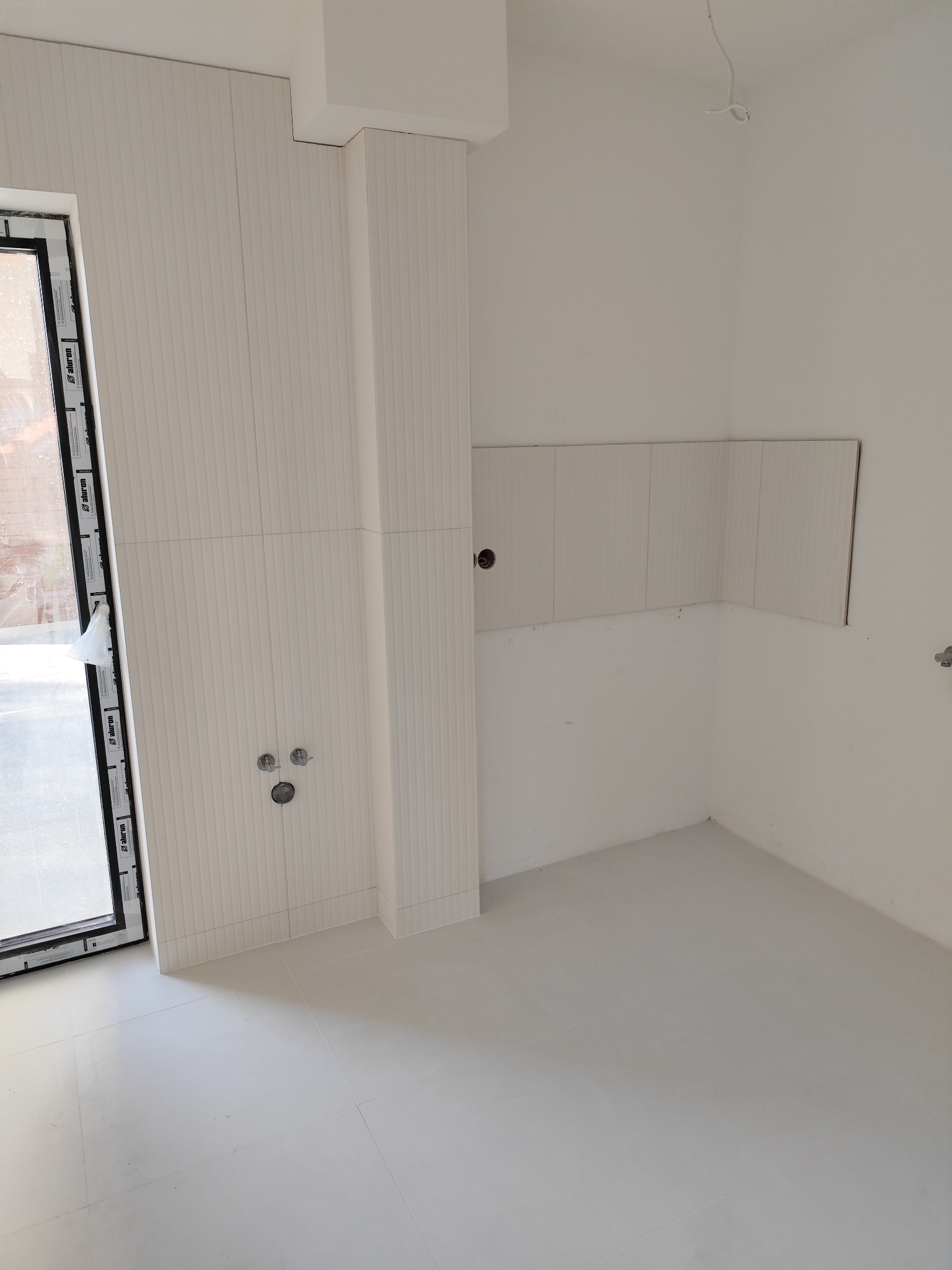 Jasne płytki ścienne z pionowym wzorem w wykończonym wnętrzu, z widocznymi przyłączami wodnymi i fragmentem okna po lewej stronie. Minimalistyczny design.